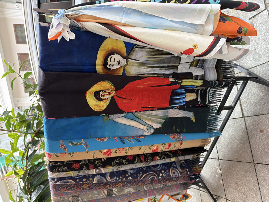 The scene depicts an outdoor retail display of textile merchandise positioned on a tiled pavement in Batumi, Georgia. A black metal rack in the foreground supports a collection of approximately ten folded or draped scarves and shawls. The items on the left side of the rack exhibit various patterns, including paisley and floral motifs, in shades of gray, navy, black, beige, and light blue. Towards the right, several scarves feature artistic prints: one light blue scarf displays a partial figure in a white garment; an adjacent black scarf shows a stylized man in a red shirt and a wide yellow hat, holding a dark object; and a dark blue scarf depicts another stylized man in a light grey shirt and a wide yellow hat, holding a light object. Additional light-colored scarves with assorted graphic patterns, including an orange floral design, are visible folded on the far right. The ground beneath the rack is composed of light grey rectangular paving tiles. In the background, a dark-colored vehicle is partially obscured behind the display, and above it, green plant foliage hangs over a light-colored building facade. No individuals are visible in the immediate foreground.