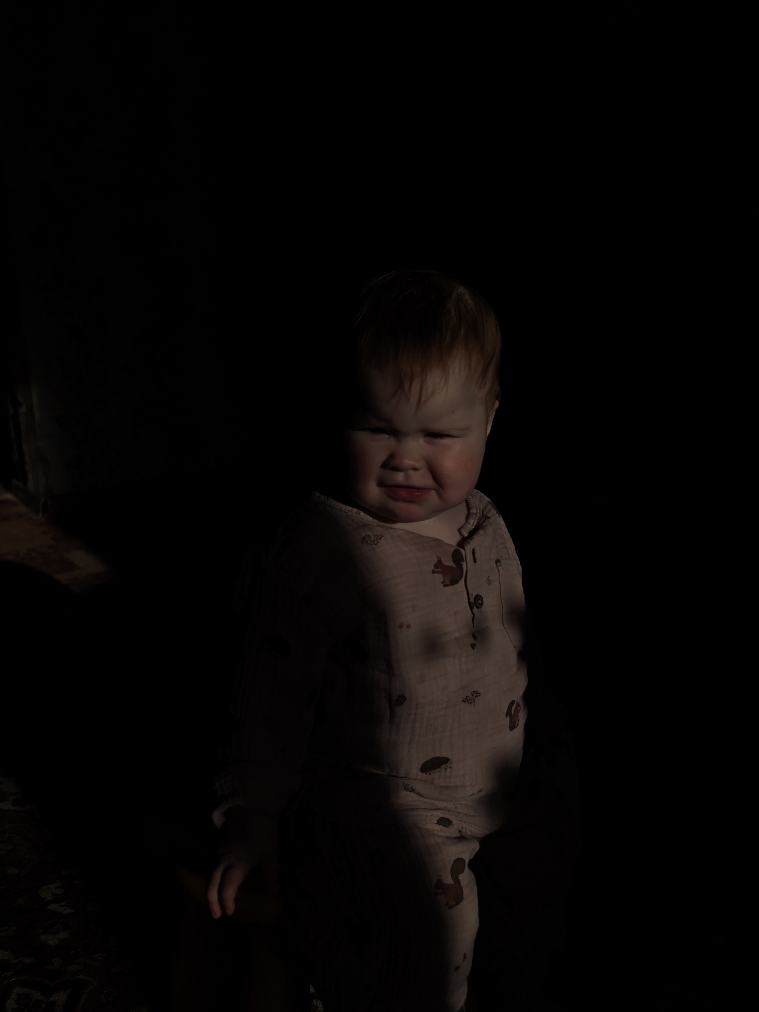 A photograph captures a young child, possibly a toddler, standing in a dimly lit environment. The child is partially illuminated by a light source from the side, highlighting their face and torso while leaving the rest of the scene shrouded in darkness. The child is wearing a light-colored garment patterned with small motifs. Their facial expression appears to be one of displeasure or discontent. Details of the surrounding environment are obscured by the darkness, although a textured surface can be discerned near the bottom of the frame, potentially a rug or carpeting. The location, Уральское, Russia, is not visually discernible from the photograph.
