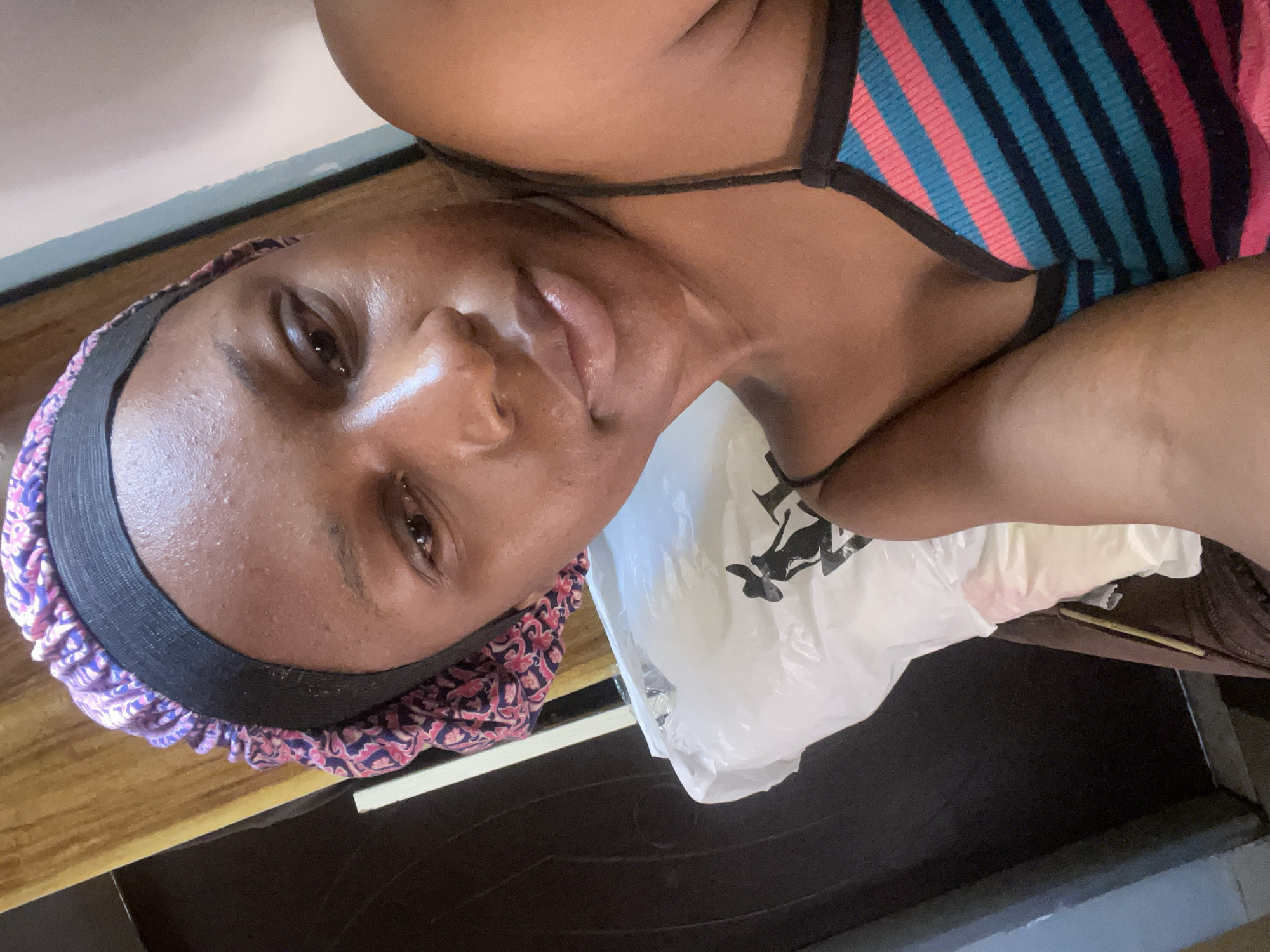 A woman is pictured in a close-up, upper-body shot, facing the camera slightly to her left. She is wearing a purple and pink patterned bonnet with a black band. Her top is a thin-strapped tank top with horizontal stripes of pink, blue, and black. She has on a light-colored face product that gives her skin a dewy appearance. To her left, a white plastic bag with a black silhouette design is visible. The background includes a dark wooden surface and a light-colored wall, suggesting an indoor setting. The image appears to be a selfie taken in a home environment. There is no information about the specific location in the city of Rafin Par, Nigeria.