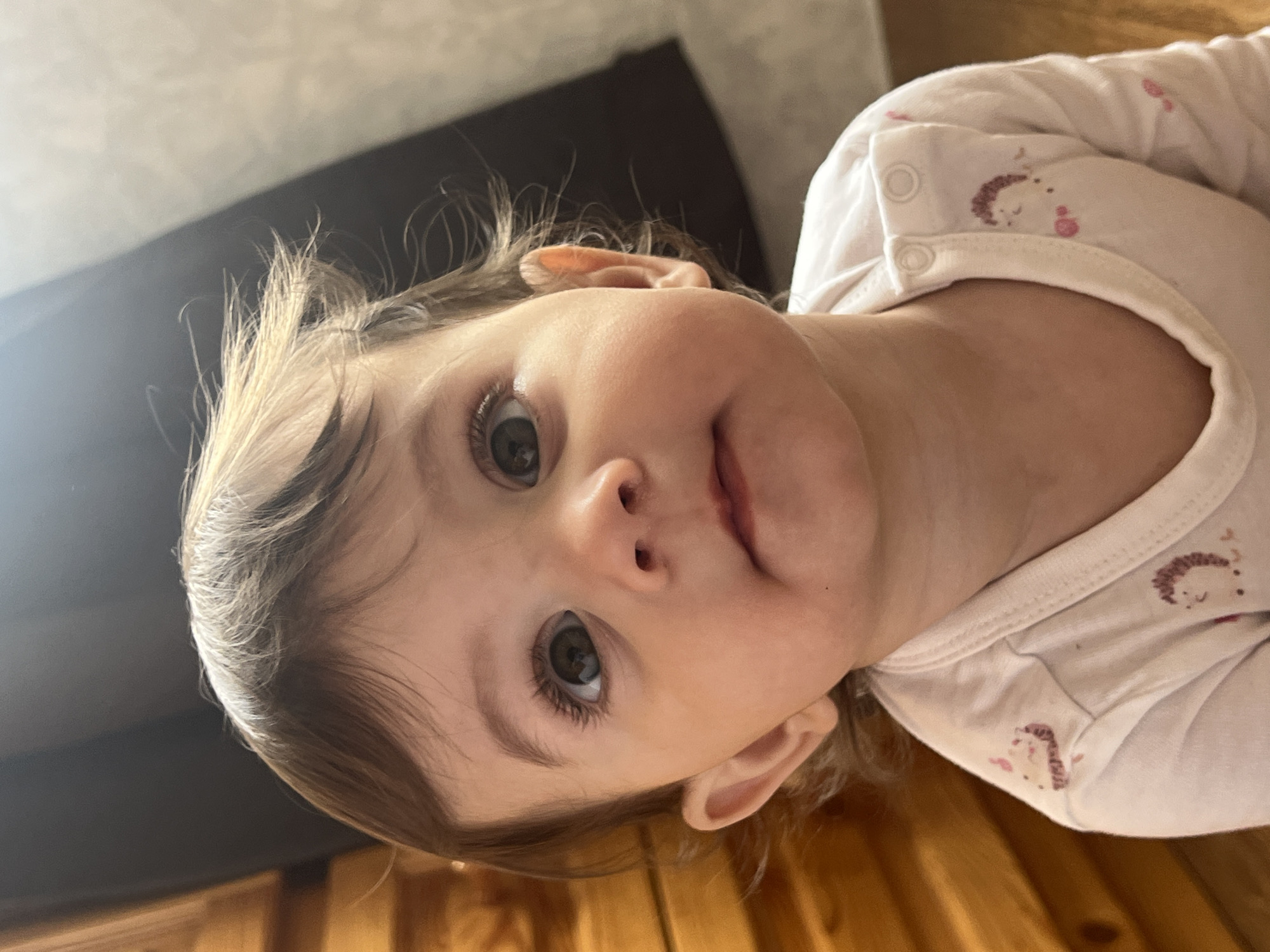 A close-up, slightly angled photograph captures a baby with light brown hair and hazel eyes. The baby is wearing a white garment adorned with a pattern of small, stylized hedgehog motifs in pink. The background includes a dark-colored surface, possibly a couch or headboard, and a wooden floor. The location of Elena, Bulgaria is not discernible within the image content.