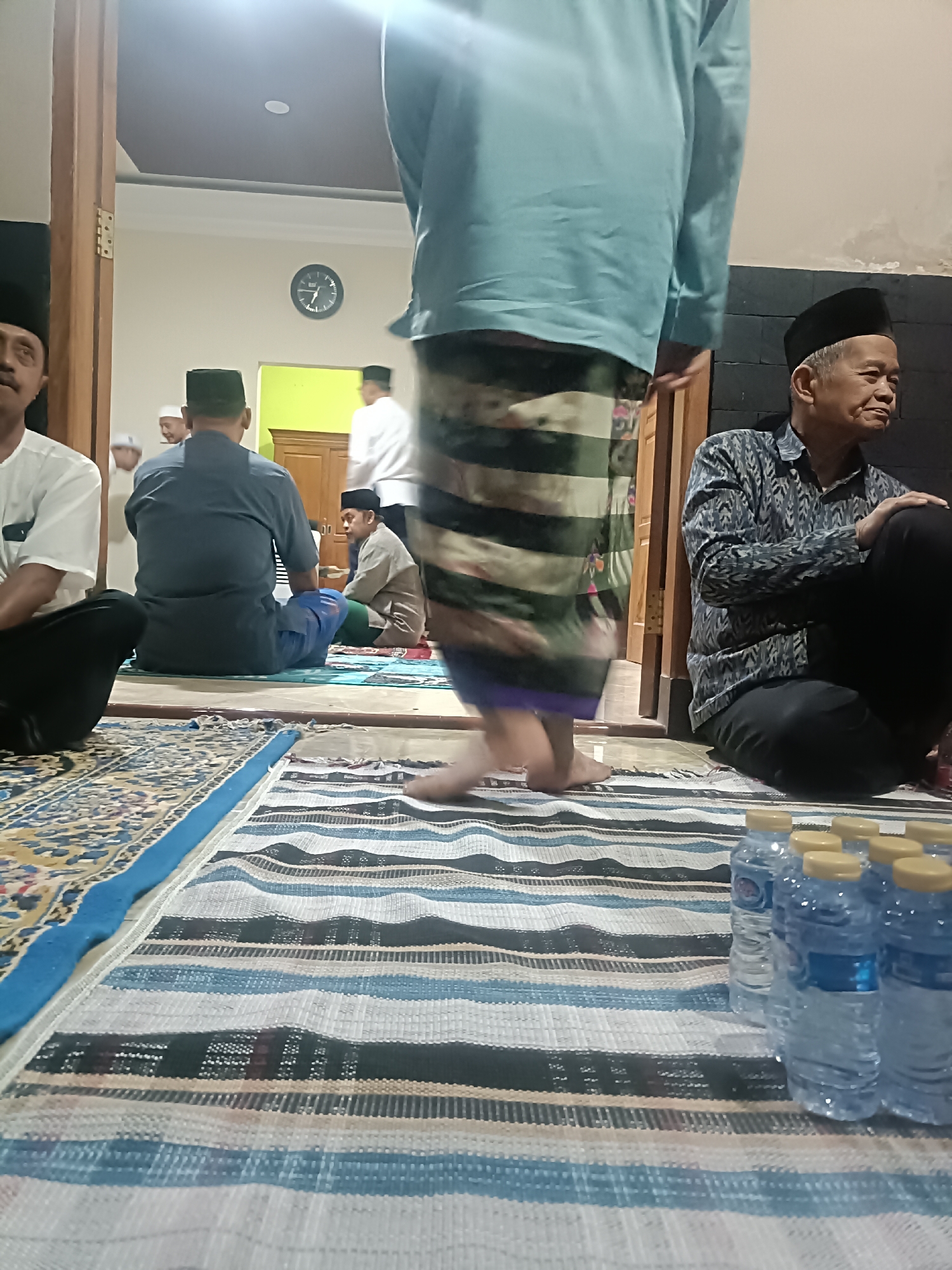 The image depicts an indoor gathering, likely in a residential or community space in Kediri City, Indonesia. Several men are present, some seated on prayer mats and others standing. In the foreground, a group of men, some wearing traditional Islamic attire including peci (caps), are seated on colorful prayer rugs. One man, in the center and slightly out of focus, is standing and appears to be wearing a light blue shirt and a sarong. To the right, an older man in a patterned shirt and black pants is seated, looking to the side. Bottles of water are arranged in the lower right corner. The environment appears to be illuminated by artificial light, and a clock is visible on the wall in the background. The overall atmosphere suggests a religious or communal gathering.