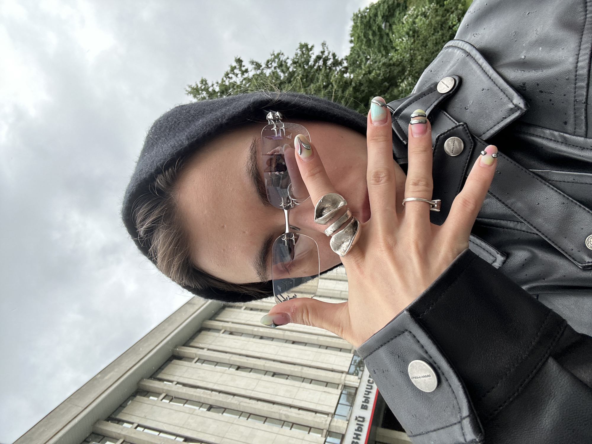 A person in a black hooded jacket and black leather jacket, wearing rimless glasses and several rings, poses for a selfie with their right hand in the foreground. The person's nails are painted with a colorful, abstract design. In the background, a large, grey building with many windows is visible, along with a cloudy sky and some trees. The building has Cyrillic text on its facade, and the person's clothing appears to be wet, suggesting it might be raining or has recently rained. The overall impression is a candid street-style portrait. Based on the architectural style and the Cyrillic text on the building, the location is likely Moscow, Russia.
