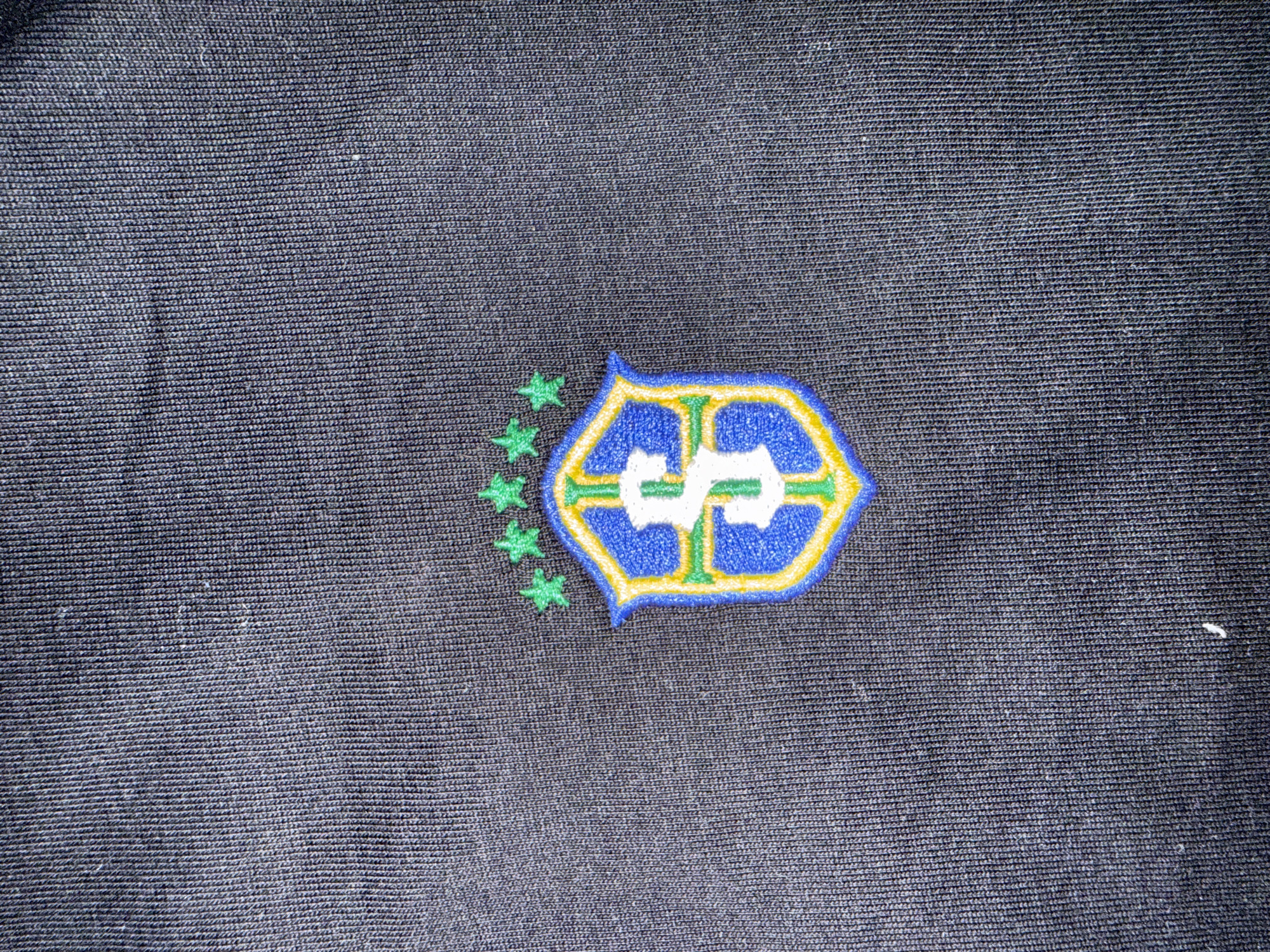 A close-up, high-angle shot shows an embroidered logo on a dark, textured fabric. The logo is shield-shaped and features a white letter "S" in the center, outlined in blue and yellow. Above the shield, there are three green stars. The fabric appears to be a ribbed knit material, possibly from a piece of clothing. The lighting is direct, casting slight shadows on the texture of the fabric. There are no people, animals, or discernible environmental features outside of the fabric and logo. This image does not provide any information about the city of Burgas, Bulgaria.