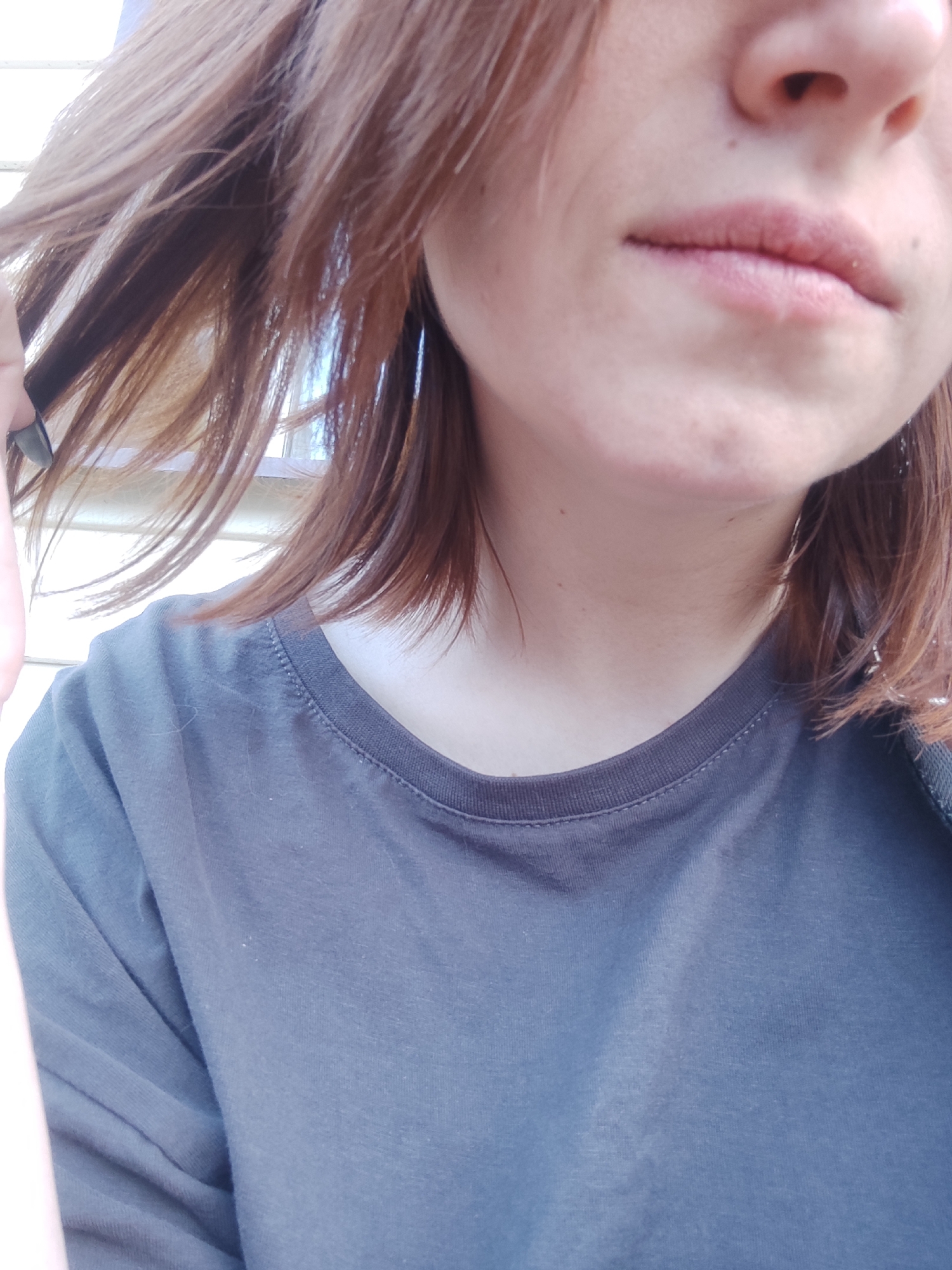 The image is a close-up, waist-up shot of a person's upper body, focusing on their face and torso. The person has reddish-brown hair that is styled in a layered cut, with some strands falling forward to obscure their face. Their mouth and chin are visible, with a light dusting of what appears to be powder or makeup on their lips. The skin on their face shows some texture and small blemishes. They are wearing a dark gray t-shirt with a visible seam around the neckline, suggesting a casual outfit.
The background is blurred, but it appears to be an outdoor setting with light-colored horizontal siding, possibly a wall or fence. The lighting suggests daytime, with a bright light source coming from the left, casting soft shadows. The overall mood is casual and unposed, with the subject seemingly interacting with their hair. There is no visible text in the image. The provided location context of Chaikovsky, Russia does not offer any specific visual cues within the image itself.