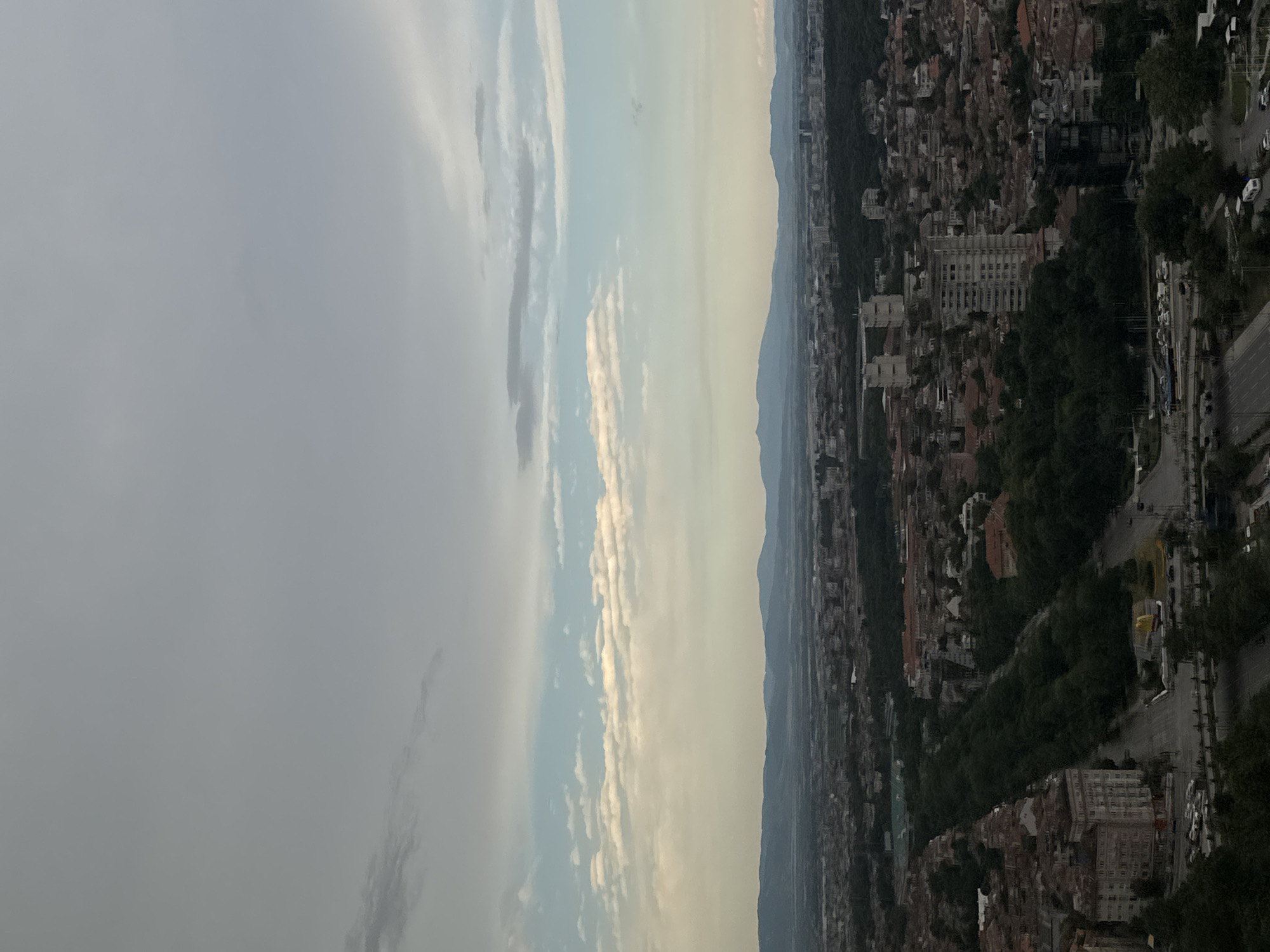 An aerial, vertical shot captures a panoramic view of Sofia, Bulgaria, under a cloudy sky with patches of blue. The scene is dominated by an expansive urban landscape stretching towards a range of distant blue mountains. In the foreground and midground, a dense collection of buildings, varying in height and architectural style, intermingle with abundant greenery, suggesting parks and tree-lined avenues. A prominent, wide, tree-lined boulevard bisects the urban sprawl, featuring roads with visible traffic and pedestrian areas. Several multi-story residential and commercial buildings are clearly discernible, including at least one taller, modern high-rise structure. The sky is a mix of grey clouds and lighter blue patches, with scattered white clouds forming horizontal bands across the mid-horizon. The overall impression is of a large, established city nestled within a natural landscape.