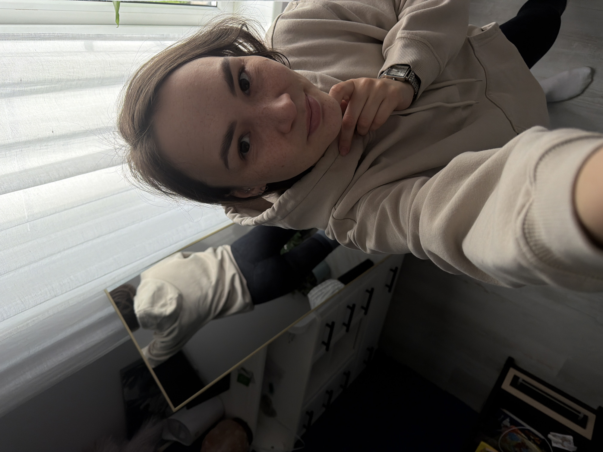 The image shows a selfie taken by a woman in what appears to be an indoor setting. She is wearing a beige hoodie and a watch. The background includes a window with white blinds and a mirror reflecting her lower body, revealing black pants and what seems to be a white sock. The reflection also shows a shelf or cabinet structure. Based on the reference to Chaikovsky, Russia, it is assumed that the photo was taken in that city, but this cannot be confirmed solely from the image.