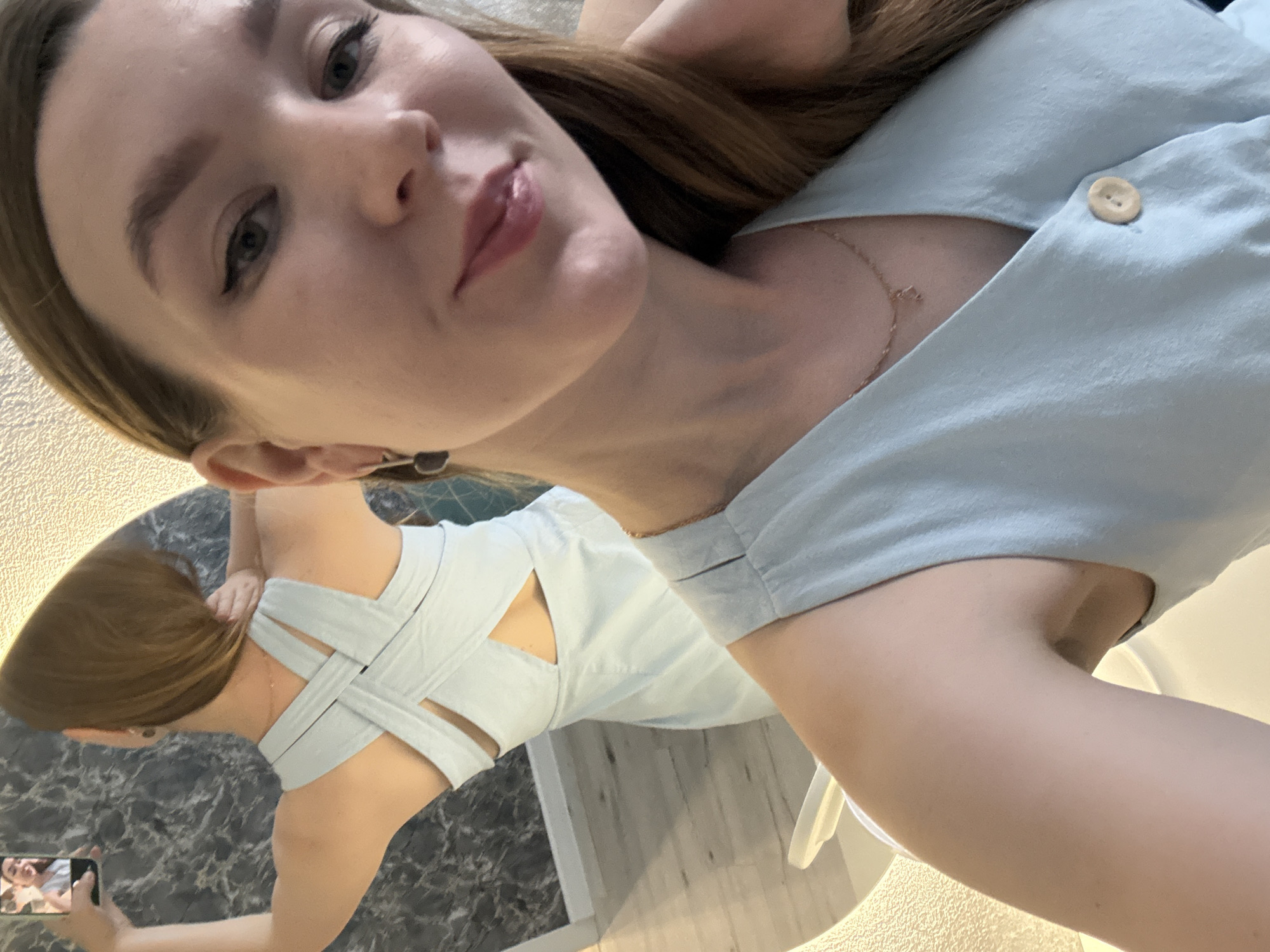 In Izhevsk, Russia, a woman takes a selfie in a light blue dress. The dress features a criss-cross back design and a V-neckline with a button closure. She is wearing a delicate gold necklace. The background shows a mirror reflecting her back and a marble-patterned wall. The scene appears to be indoors, possibly in a home or fitting room.