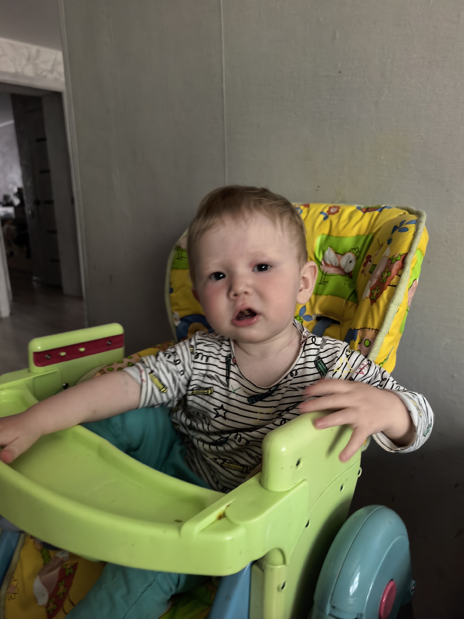 A toddler sits in a green and yellow high chair. The child wears a long-sleeved, striped shirt with various objects printed on it. The high chair's seat is lined with blue fabric, and the child's pants appear to be teal. The room has grey walls. The location is Sterlitamak, Russia.