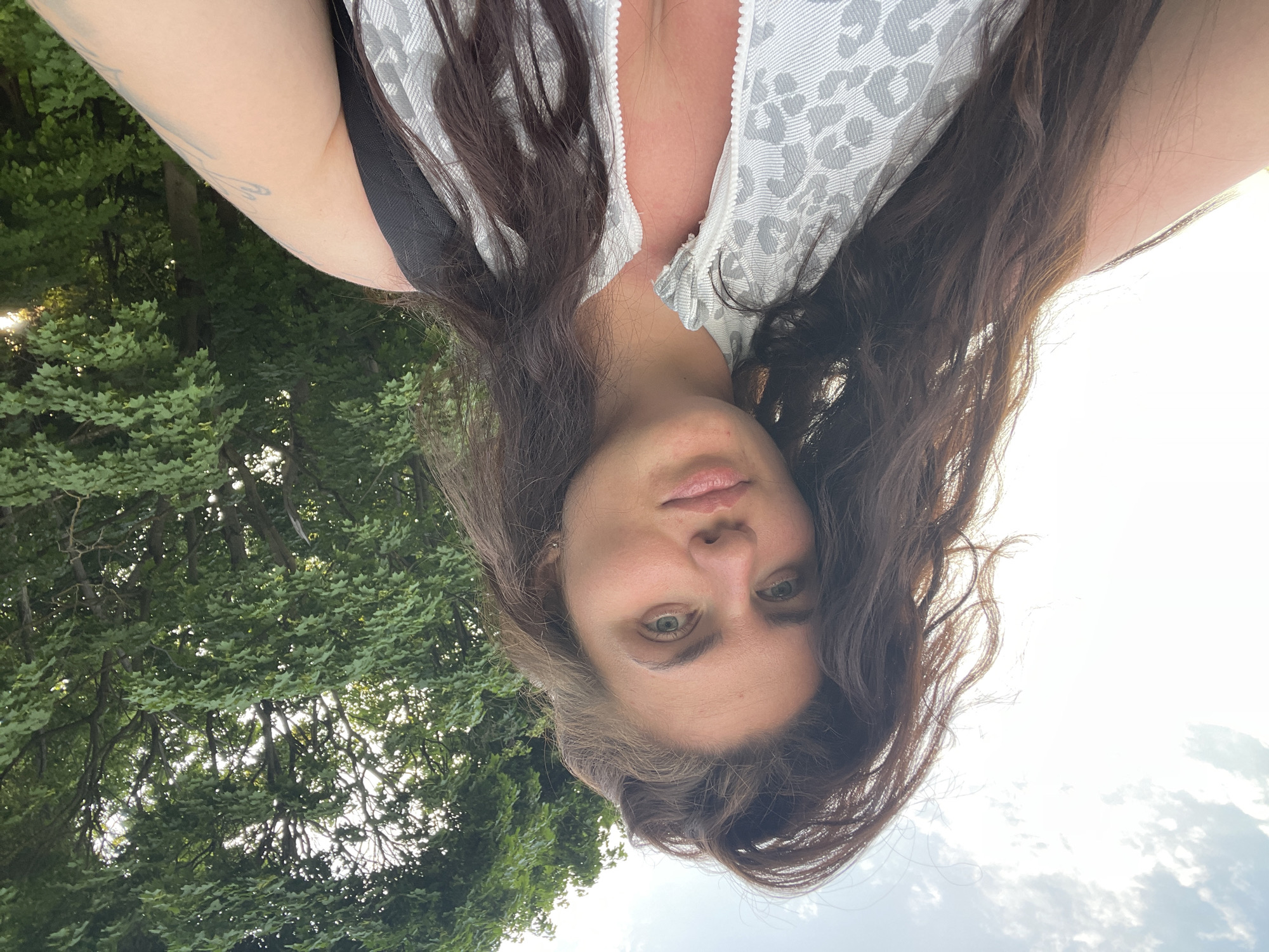 A selfie shot outdoors depicts a woman with long brown hair, green eyes, and fair skin. She is wearing a light-colored top with a leopard print and a black tank top underneath. The background includes green trees and a bright sky. There is no indication of the city of Ostrov, Russia.