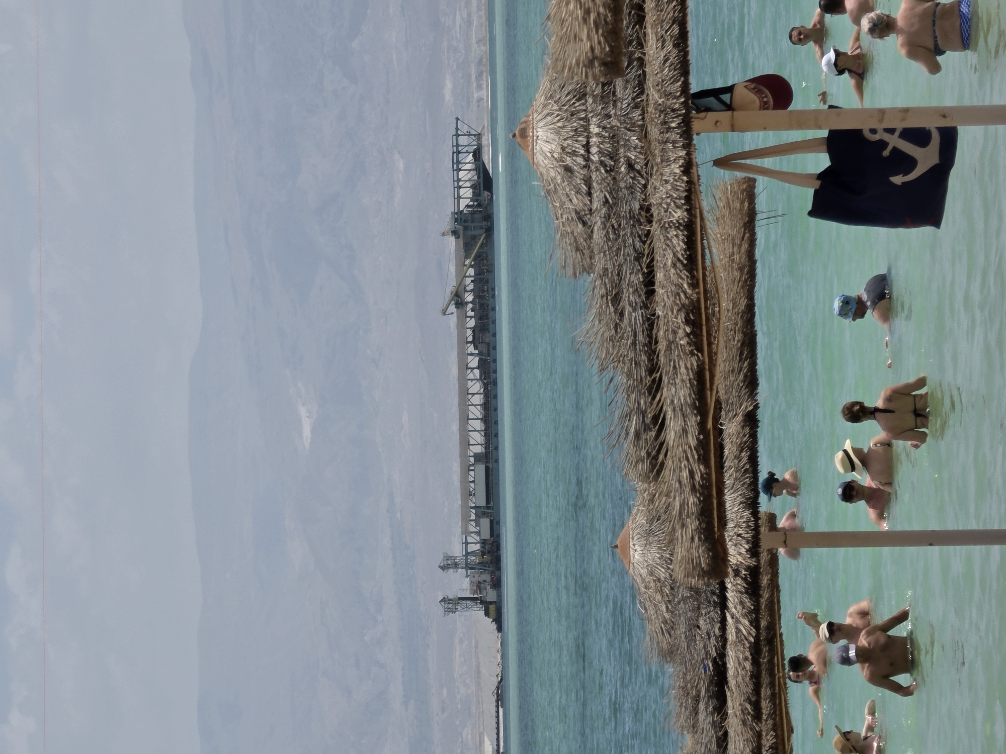 The image depicts a scene at the Dead Sea in the Tamar Regional Council, Israel. A long pier-like structure with industrial equipment is visible along the shoreline. In the foreground, there are several straw umbrellas, typical of beach setups. A group of people are enjoying the Dead Sea, some wading in the water, some swimming. A tote bag with an anchor is hanging from one of the umbrella poles. The water is turquoise in color, and the sky is slightly hazy. The background features a mountainous landscape.