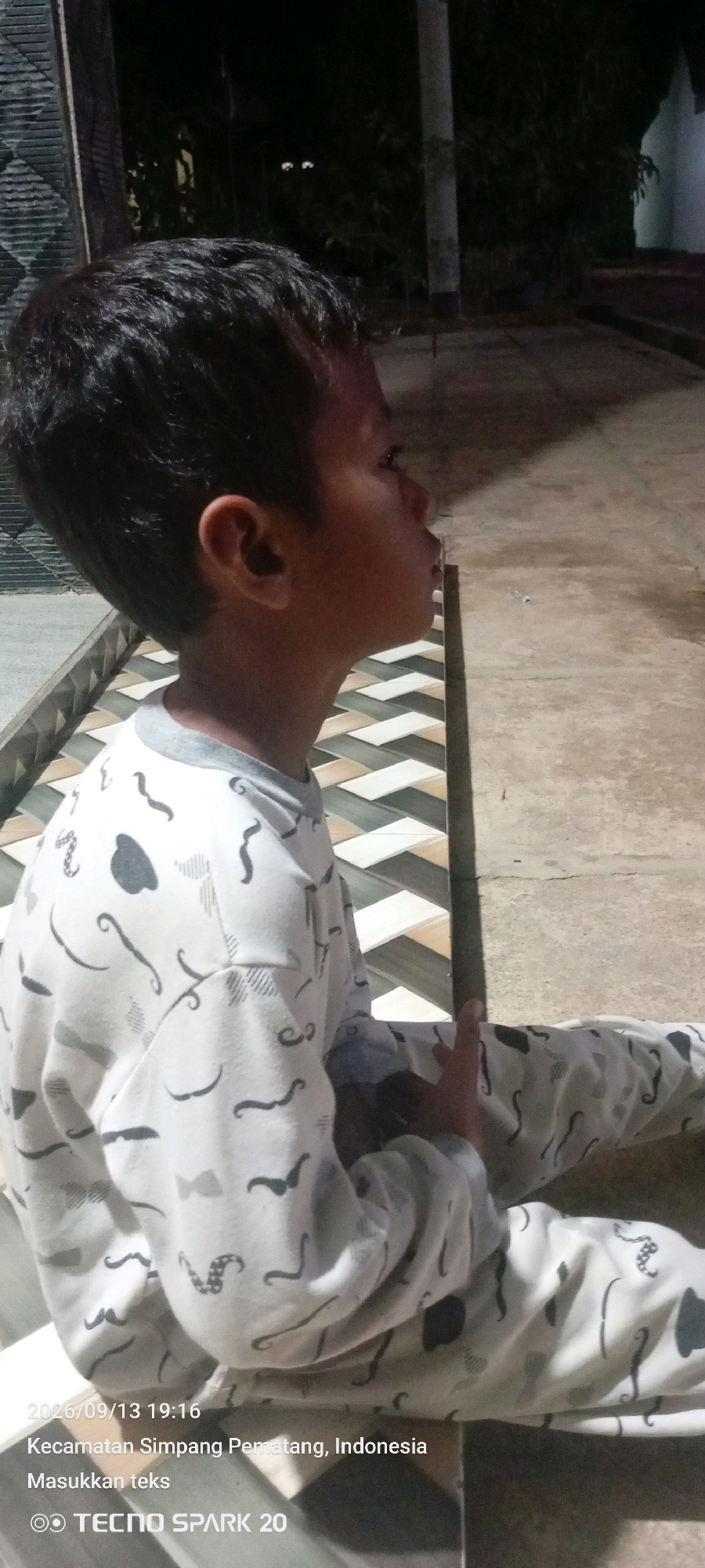A young boy with dark hair, wearing a white long-sleeved shirt with a pattern of mustaches and other black designs, is sitting on the ground in Simpang Pematang, Indonesia. He is positioned in profile, looking to the right. The ground beneath him is a patterned tiled surface, transitioning to concrete. The background is dark, with hints of foliage and structures, suggesting an outdoor setting at dusk or night. The timestamp "2026/09/13 19:16" is overlaid on the image, along with the text "Kecamatan Simpang Pematang, Indonesia" and "Masukkan teks," and the device branding "TECNO SPARK 20".