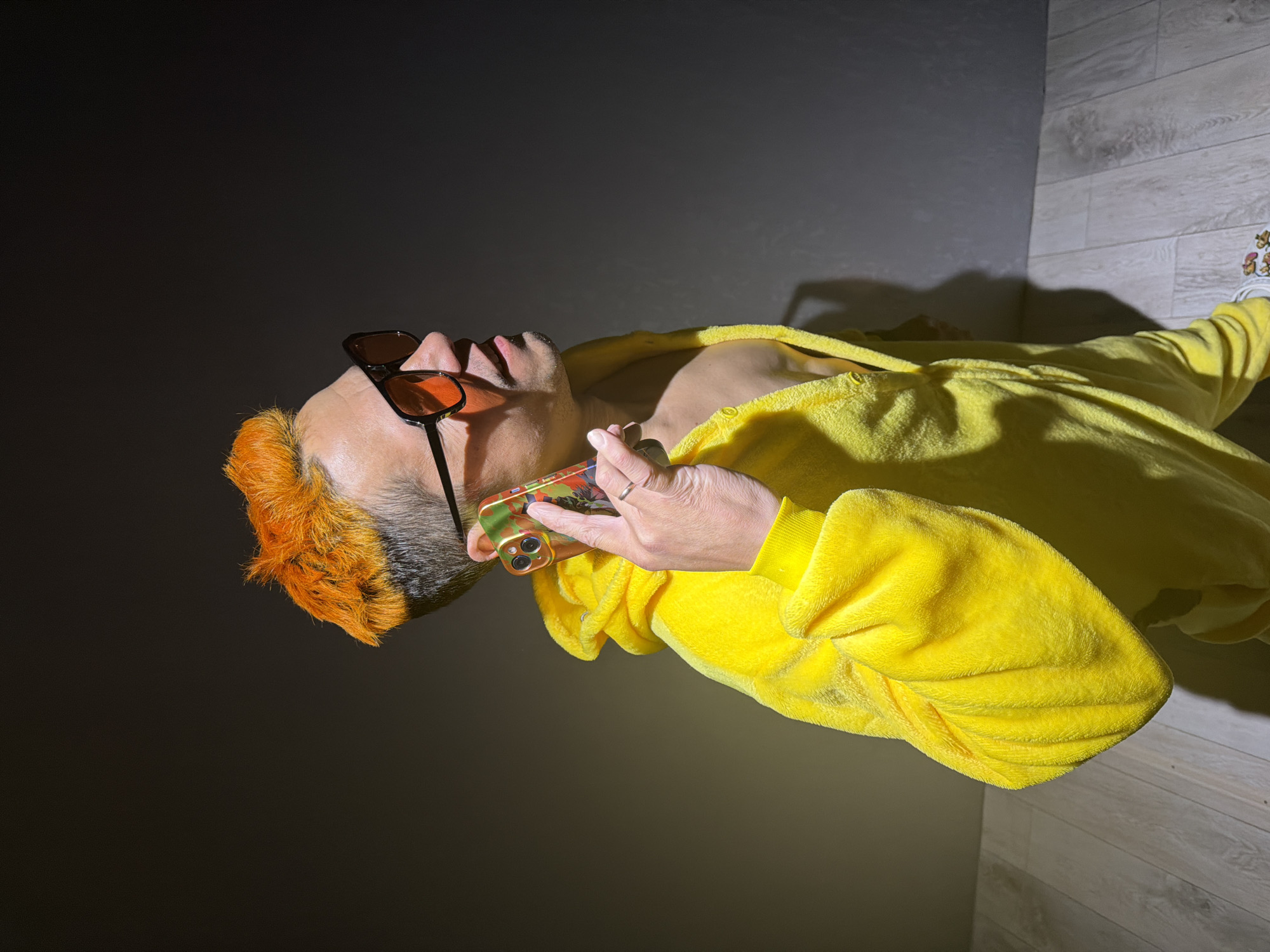 The photograph shows a person wearing a yellow robe and sunglasses, holding a smartphone to their ear. The person has short, orange-dyed hair. The background is a combination of a dark wall and a lighter wood-paneled wall or floor. The subject is standing indoors. There is no indication of the city of Chaikovsky, Russia.
