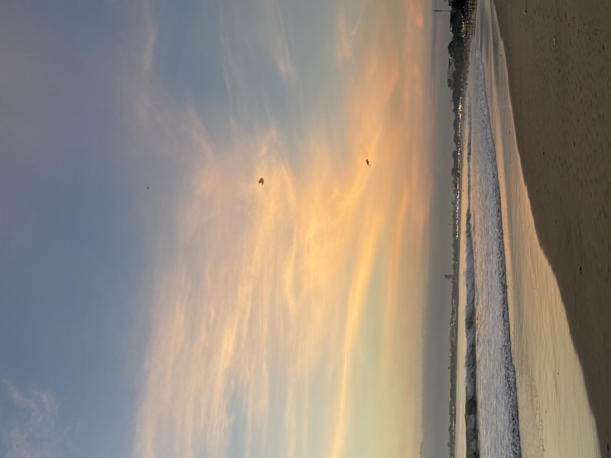 The image captures a beach scene at sunset in Kuta Selatan, Indonesia. The sky transitions from blue to orange with wispy clouds. Birds are visible in the sky. Waves are gently lapping on the shore, which is a light brown. The edge of the beach has buildings and lights that line the coast.