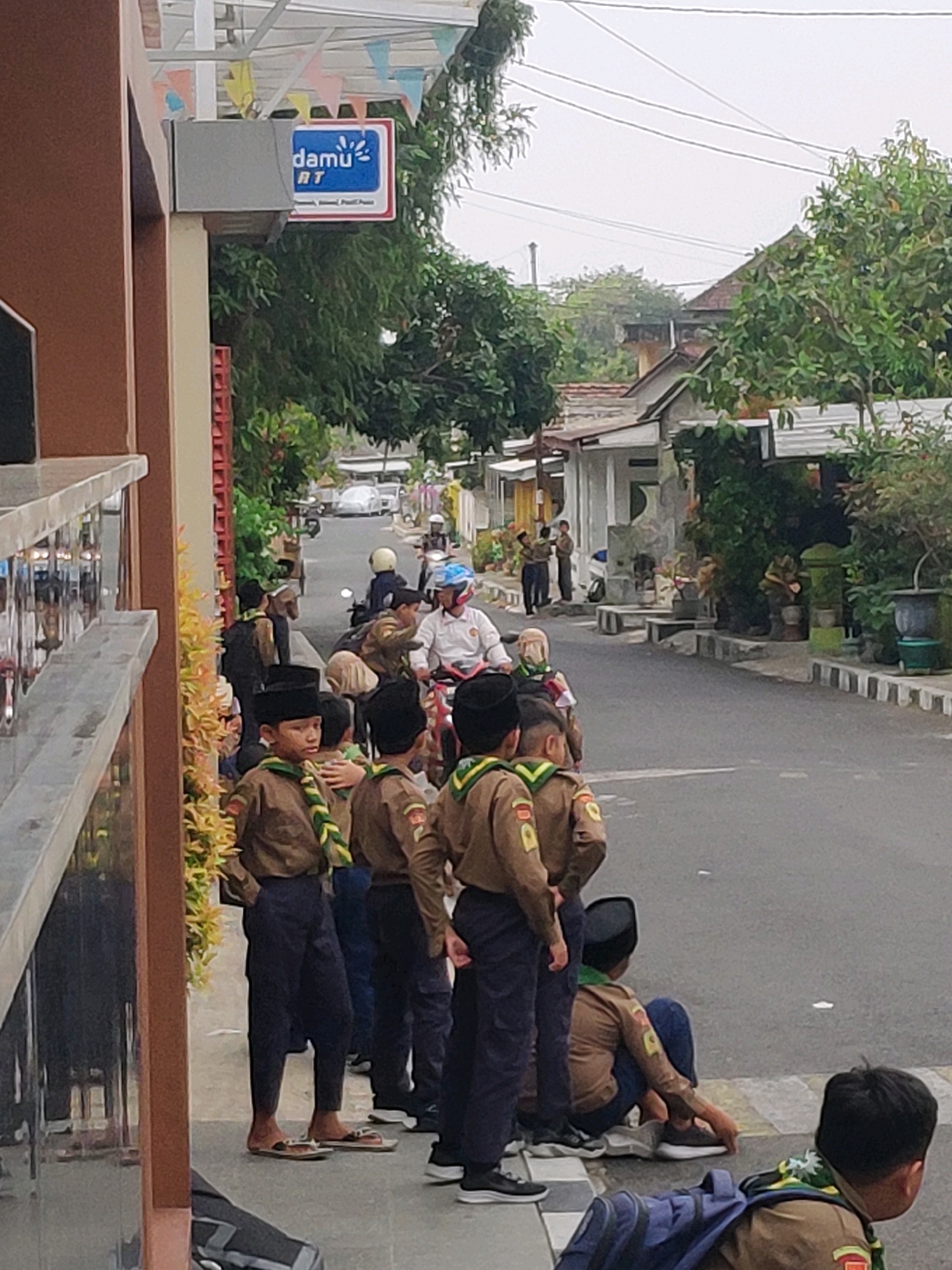 The image depicts a street scene in Magetan, Indonesia. A group of young people, appearing to be scouts based on their uniforms, are gathered on the side of a road. They are wearing brown shirts with green and yellow neckerchiefs and dark pants or shorts. Some are standing, while others are sitting on the curb or the ground. One individual in the foreground is wearing a black cap and a scout uniform. Further down the street, a person on a motorcycle is visible, wearing a white shirt and a helmet. Buildings line the street, and trees and foliage are present, suggesting a residential or commercial area. A sign with the text "damu AT" is mounted on a pole near the building on the left. The sky is overcast.