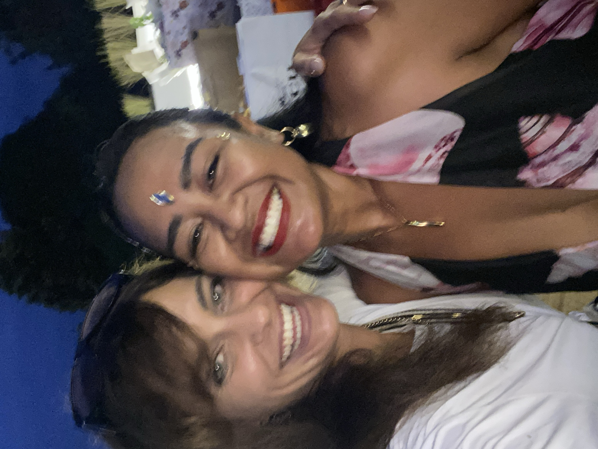 This media file is a selfie featuring two women, likely friends, enjoying themselves. The woman in the foreground has long, dark brown hair and is wearing sunglasses pushed up onto her head. She is smiling widely, revealing her teeth. She is wearing a white top. The second woman, positioned behind and slightly above the first, has darker skin and is also smiling. She has dark hair and is wearing a black top with a floral pattern. A sparkling blue jewel or sticker is visible on her forehead, and she has gold earrings and a necklace. The overall impression is of a happy and celebratory atmosphere.
The image appears to have been taken outdoors, possibly at night or in a dimly lit setting, given the overall darkness and the visible blue of what could be a sky or water. The presence of what looks like foliage or decorations in the background suggests a festive event or social gathering. The location context of Nusa Penida, Indonesia, implies a tropical setting, and the warm lighting and relaxed attire of the subjects are consistent with this. The emotion conveyed is one of joy, connection, and fun. There is no visible text in the image.