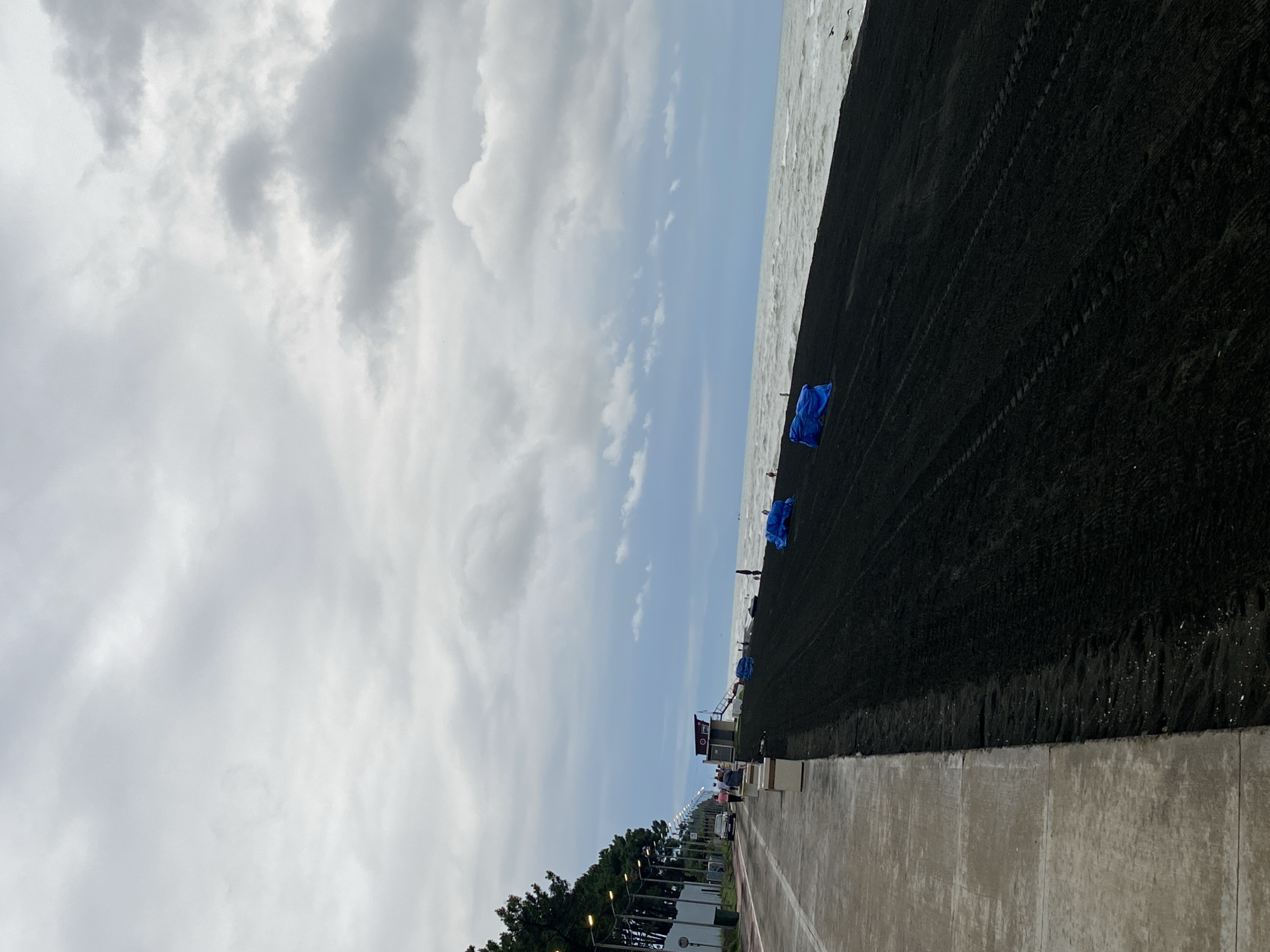 The image depicts the coastline of Ureki, Georgia, a town known for its magnetic black sand beaches. The scene is dominated by the dark, textured sand stretching from the foreground towards the sea. The sky is overcast with heavy, grey clouds, creating a muted light that diffuses across the landscape. The sea is moderately rough, with visible waves and whitecaps reaching the shore.
On the left side of the image, a paved promenade runs parallel to the beach, separated by a low wall and a row of streetlights. Several individuals are visible on the promenade, with some walking and others seated. A car is parked near the promenade. Adjacent to the promenade, a line of tall, dark green trees forms a natural barrier.
Along the edge of the promenade and bordering the black sand, there are white, rectangular benches or planters. Further along the beach, closer to the sea, are several large, bright blue tarps, suggesting some form of construction or preparation work is underway. A small, elevated structure, possibly a lifeguard station or information booth, is situated near the water's edge. The overall impression is one of a coastal area undergoing development or maintenance, under a dramatic, cloudy sky.