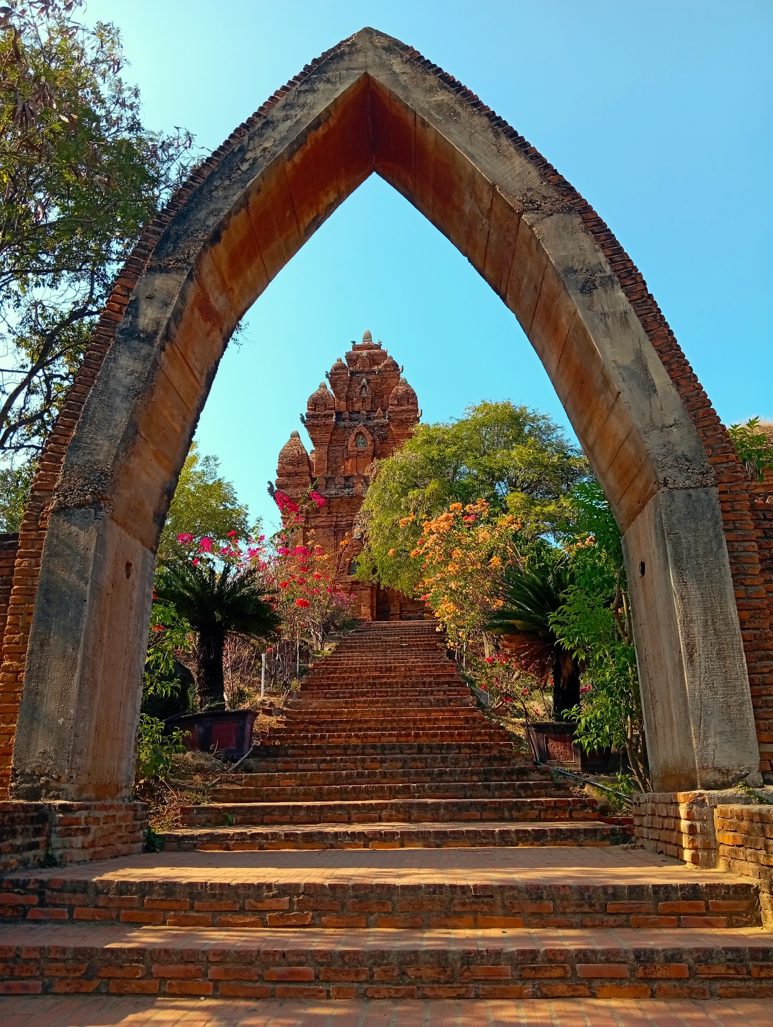 This vertical shot captures a grand, ancient-looking archway made of weathered red brick and concrete, framing a long staircase that ascends towards a multi-tiered temple. The archway, with its worn texture and some exposed brickwork, spans the width of the frame in the foreground, creating a natural gateway to the scene beyond.
The red brick staircase dominates the central view, leading the eye upwards. On either side of the stairs, lush green foliage, palm trees, and vibrant flowering bushes in shades of pink, orange, and red add splashes of color and life to the historical setting.
At the top of the stairs, partially obscured by the archway and surrounding trees, stands a distinctive brick temple or tower structure. It features multiple ornate tiers and intricate architectural details characteristic of ancient Southeast Asian, possibly Champa, design. The structure appears to be made of the same reddish-brown brick as the stairs and archway, suggesting a cohesive architectural style.
The sky above is a clear, brilliant blue, indicating fair weather. Bright sunlight illuminates the scene, casting soft shadows across the brick steps, suggesting a late morning or early afternoon setting. No people are visible, emphasizing the serene and historic atmosphere of the site. There is no visible text in the image. The location is Phan Rang – Tháp Chàm, Vietnam, consistent with the architectural style.