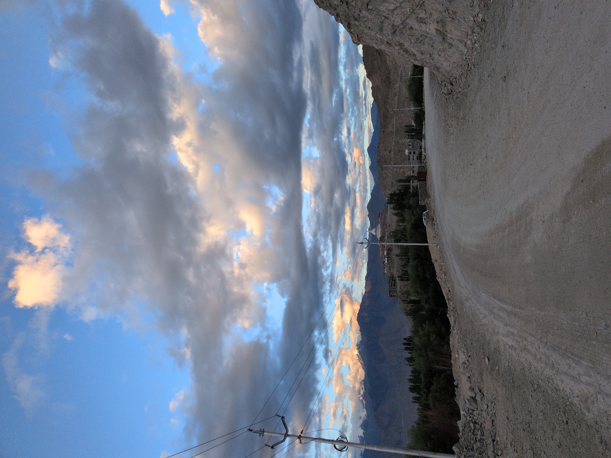 An exterior, eye-level, vertical, medium shot captures a dirt road curving towards the right, leading toward a settlement nestled in a valley beneath snow-capped mountains under a cloudy sky. The road is a light brown dirt with visible gravel and is surrounded by rocky terrain. To the left of the road, a utility pole with several wires stands prominently. In the background, a cluster of buildings with white roofs, including a prominent stupa, sits on a hill. The mountains in the distance are dark with snow-covered peaks. The sky is a mix of blue and gray clouds, with some areas illuminated by the setting sun, casting warm hues on the clouds. The scene appears to be located in Leh, India, given the landscape and architecture. No people are visible in the image.