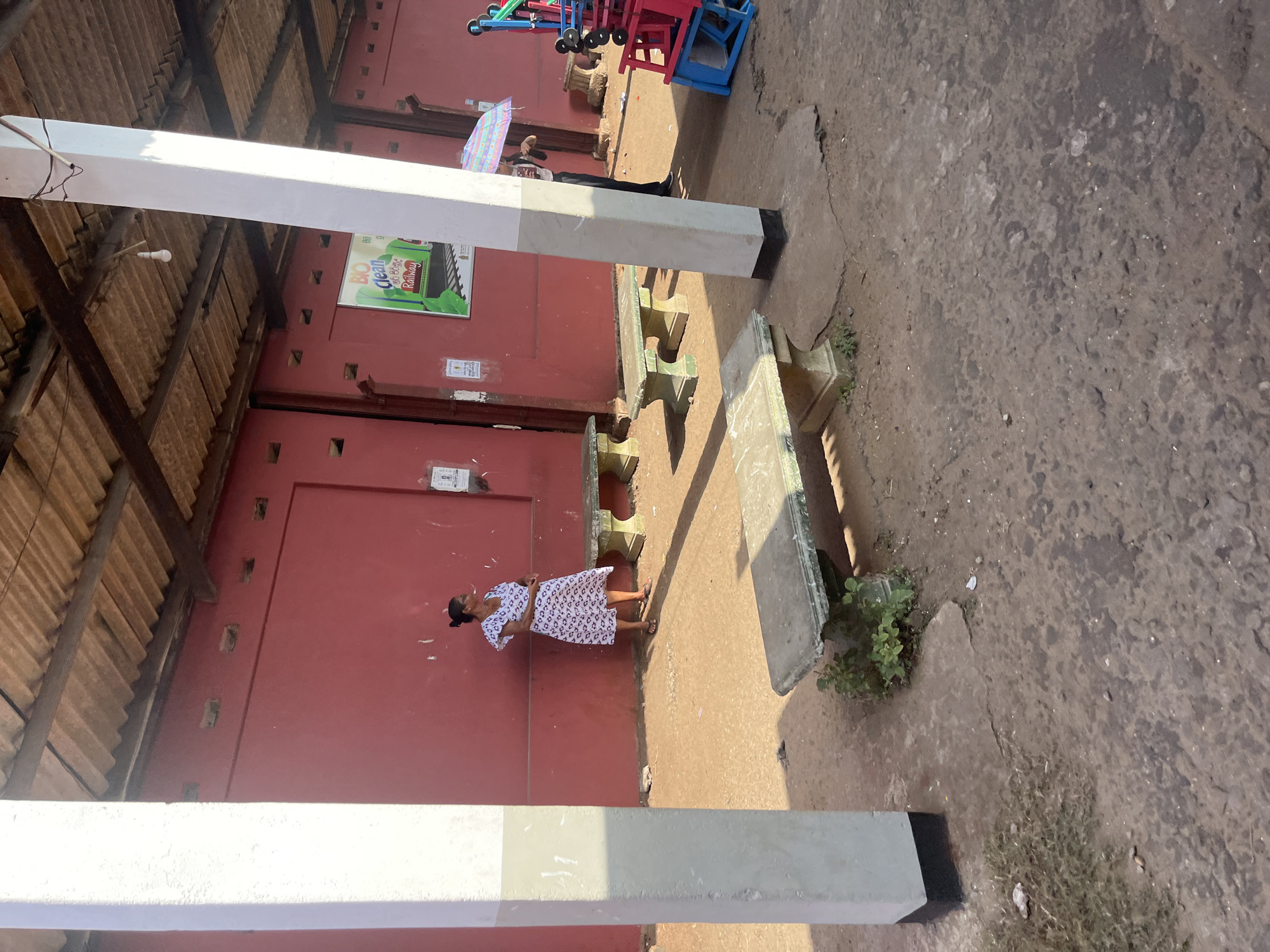 The image depicts an outdoor scene at a bus stand in Moratuwa, Sri Lanka. A woman in a patterned dress stands by a red wall, facing left and looking away from the camera. Several concrete benches are present in the foreground and midground, some with visible weathering and small plants growing around them. In the background, a sign with the text "BIO Clean Railway" is visible on the red wall. To the right, a person holds a colorful umbrella, and some exercise equipment is stacked. The ground is a mixture of dirt and concrete. The architecture suggests an open-air structure with a corrugated roof supported by wooden beams and prominent white pillars. The overall impression is a functional, somewhat worn, public space.