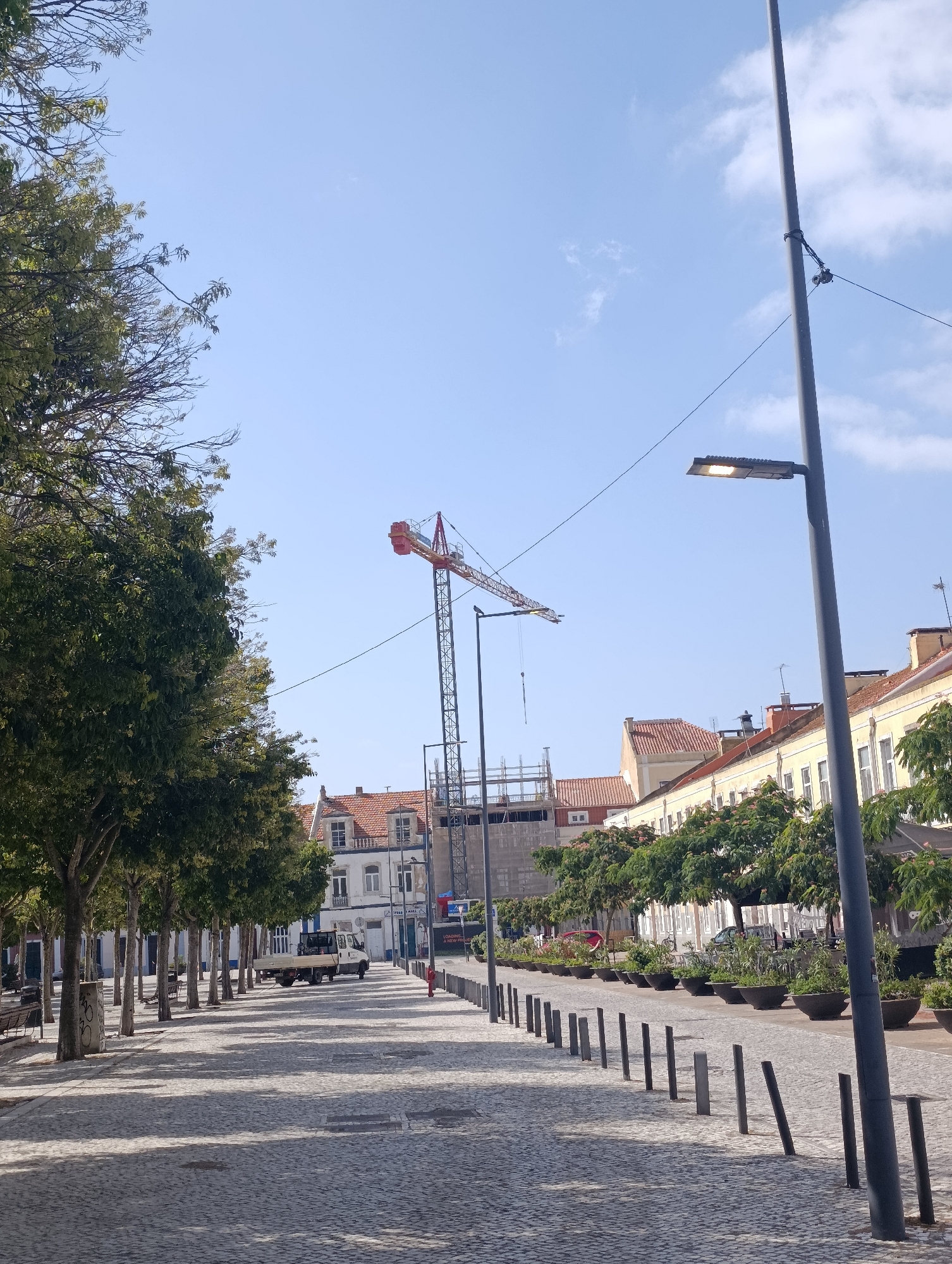 The image depicts a street scene in Setúbal, Portugal, featuring a paved walkway lined with trees and buildings. A prominent construction crane is visible in the background, indicating ongoing development. The street is paved with cobblestones, and there are several lampposts and bollards along the walkway. A white truck is parked on the street, and there are potted plants lining the side of the road. The sky is clear with some scattered clouds. The buildings appear to be a mix of residential and commercial properties, with tiled roofs and visible windows. The trees are full of green leaves, suggesting a temperate season.