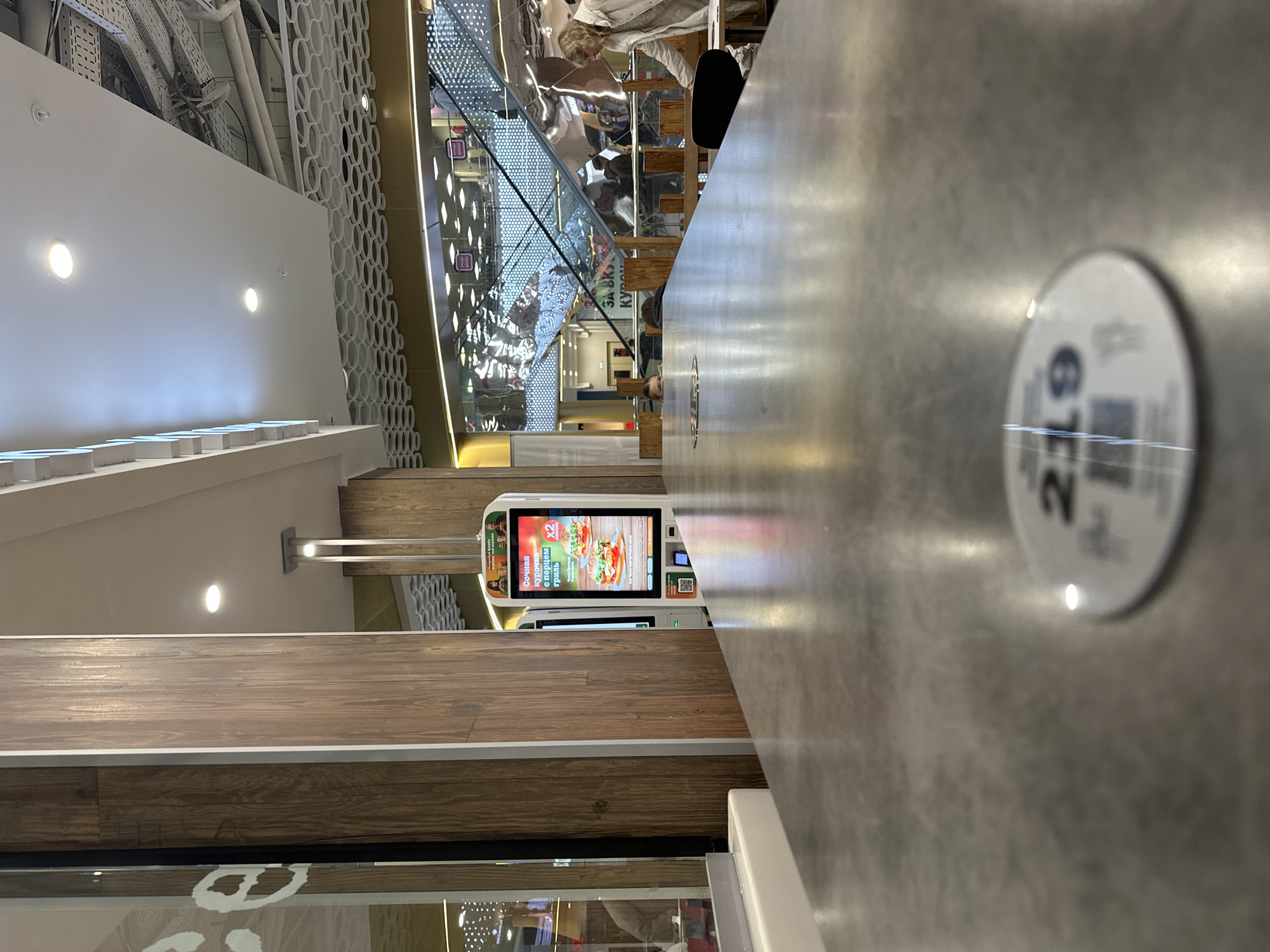 The image captures an indoor scene in Kazan, Russia, likely within a shopping mall or food court. A self-service kiosk with a brightly lit digital menu screen is visible, showcasing food items and advertising "X2" deals. The kiosk is positioned near a wooden-paneled wall with white trim. To the right, a gray countertop table extends into the foreground, marked with a small round sticker bearing the number "26." A blurred view through a glass partition reveals an escalator and other shoppers. The overall environment appears modern and commercial, with an emphasis on self-ordering food.