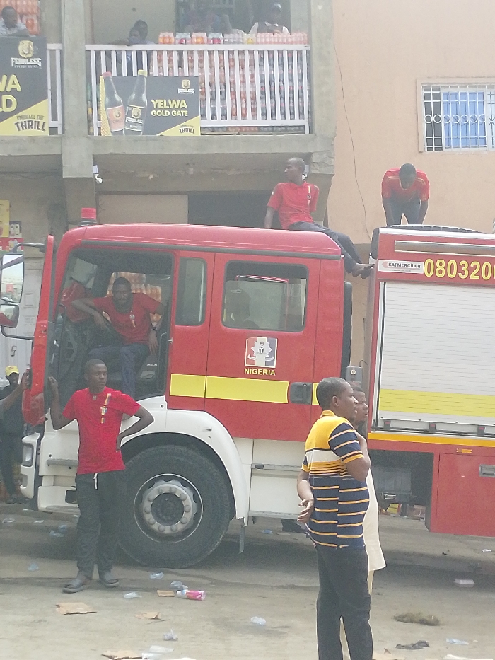 The scene depicts a red fire truck, likely a MAN chassis, parked on a paved surface in Fagge C, Nigeria. The truck bears the inscription "NIGERIA" on its side and "KATMERCILER 0803200" on the rear section. Several individuals are present: four men in red t-shirts are positioned on and around the fire truck. One male sits atop the cab, facing right. Another male leans over the rear compartment. A third male stands by the driver's side front wheel, looking towards the right, with a hand on his hip and multiple badges/medals on his chest. A fourth male leans out of the driver's side door, looking forward. Two other males are visible in the foreground: one in a horizontally striped yellow and blue t-shirt and dark pants, and another partially obscured figure. The immediate ground area is littered with various debris, including plastic bottles and paper. Behind the fire truck, a multi-story building is visible. Its second floor features a balcony filled with stacks of bottled beverages. Advertising banners for "PEERLESS ENERGY DRINK" and "YELWA GOLD GATE" are affixed to the building's facade. Additional blurred figures are present on the balcony behind the stored beverages.