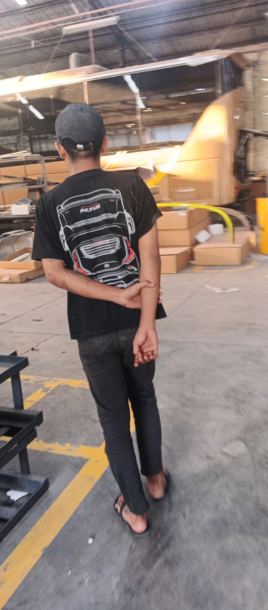 A person wearing a black t-shirt with a graphic of a car's rear end and dark pants stands with their back to the camera, facing a large, light-colored vehicle. They are also wearing flip-flops. The person's arms are behind their back, and their left arm is crossed over their right. They are wearing a dark baseball cap. The environment appears to be an indoor industrial space, possibly a warehouse or factory, with concrete flooring marked with yellow lines. Cardboard boxes are stacked in the background. The overall lighting is somewhat dim, with fluorescent lights visible on the ceiling. The image was captured in Banjararum, Indonesia.