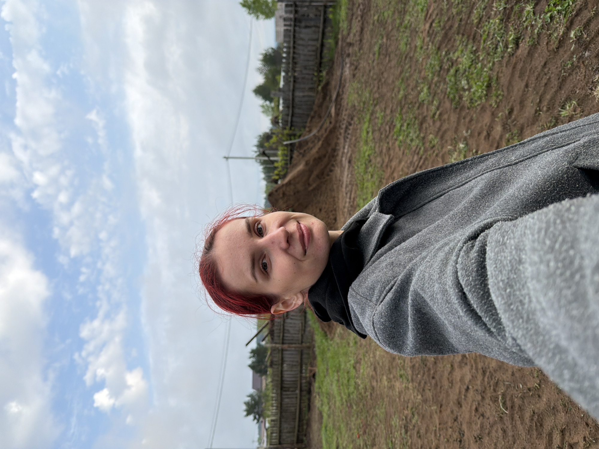 A medium shot depicts a person with fair skin and red hair, angled slightly left, with a landscape behind them. The person is wearing a gray hooded sweatshirt. The background shows a rural landscape consisting of brown soil sparsely covered with green vegetation. A wooden fence runs horizontally across the midground of the image. Beyond the fence, glimpses of buildings and trees suggest a residential area. The sky above is overcast with scattered clouds. The location is in the city of Chaikovsky, Russia.
