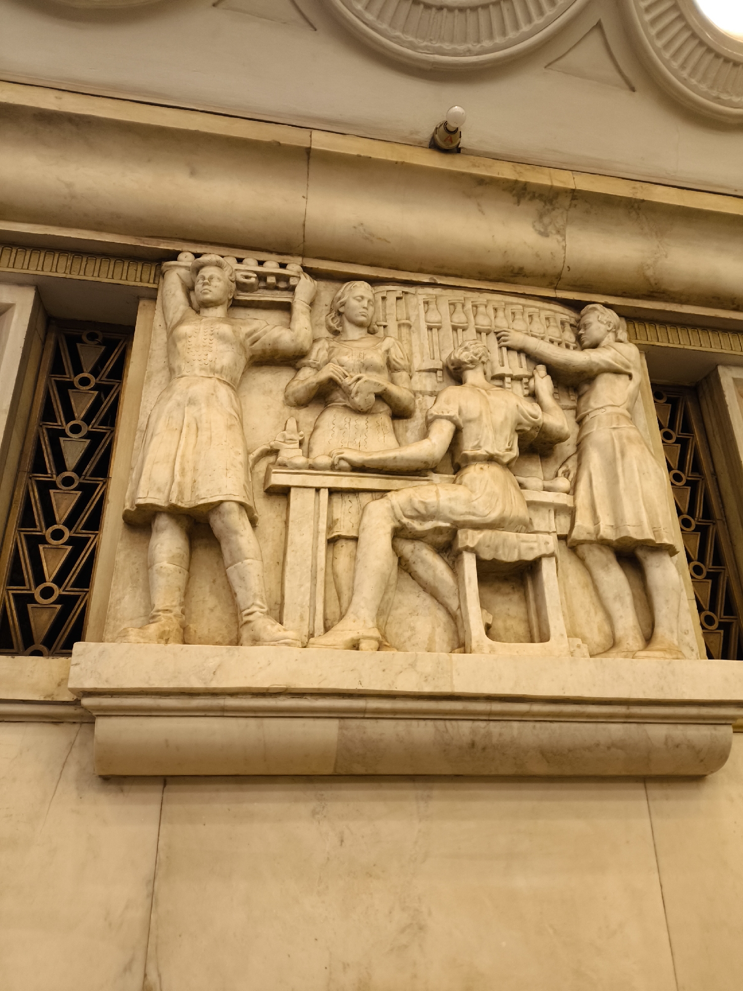 Captured at an indeterminate location within the city of Unknown, Unknown, the image showcases a marble relief carving mounted on a light-colored stone wall. The sculpture depicts a group of four figures, possibly women, dressed in what appear to be early 20th-century attire. One woman stands on the left with her arms raised, as if holding something above. Another stands beside her, gazing at an object in her hands. In the center, a woman sits at a table, while a fourth figure stands behind her, seemingly adjusting or working on her hair. Behind these figures, one can discern a background suggestive of shelves or a rack containing numerous bottles or containers. To the left and right of the relief are dark ornamental grilles. Positioned above the sculpture is a section of decorative molding and a light fixture.