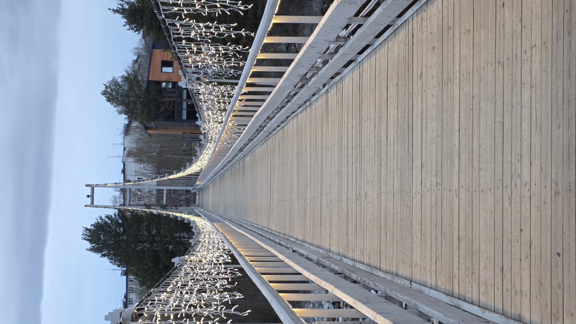 The image shows a long, wooden pedestrian suspension bridge in Kandalaksha, Russia. The bridge deck is made of vertical wooden planks. White, ladder-style railings flank both sides of the bridge. Decorations of small, white lights are strung along the railings. In the background, buildings and trees are visible. The sky is light blue and overcast. The perspective of the image suggests it was taken from the bridge, looking straight down its length.