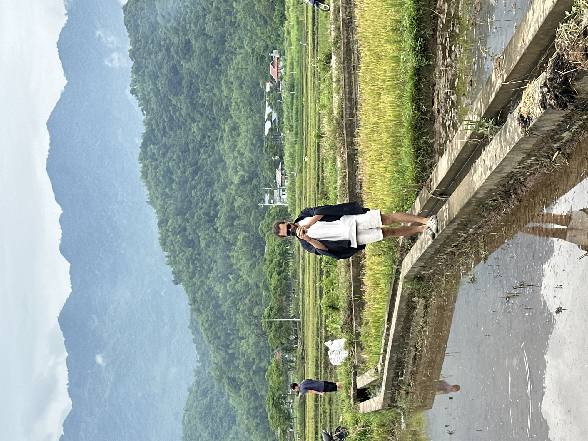 In Mai Chau, Vietnam, a man with sunglasses stands on a concrete path dividing a flooded rice paddy from a mature rice field. He is wearing a white t-shirt, white shorts, and a dark open jacket, and he holds a smartphone to his face, appearing to take a photograph or video. In the background, a second person, wearing a dark blue t-shirt and black shorts, stands facing away from the camera. Beyond the rice fields, a line of trees separates the agricultural land from a cluster of buildings. The landscape is framed by lush green mountains under a cloudy sky.