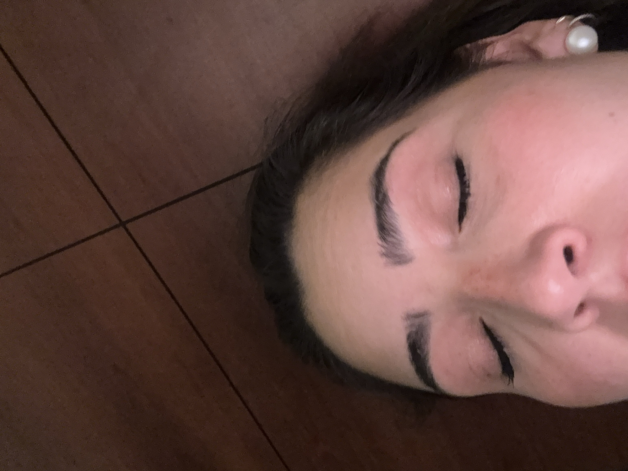 A close-up, angled view of a person lying down with their eyes closed. The person has dark hair and defined eyebrows. They are wearing a pearl earring. The surface beneath the person appears to be tiled with dark brown, square tiles. The location is unable to be determined.