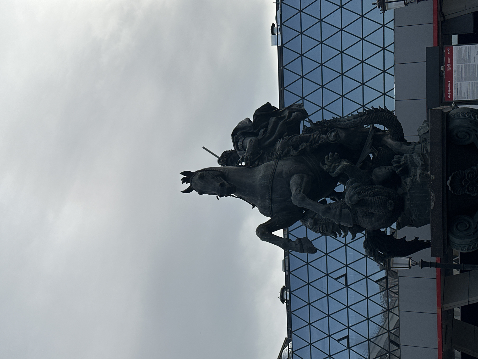 The image shows a statue of Saint George slaying the dragon in Moscow, Russia. The statue depicts a mounted knight, presumably Saint George, piercing a dragon with a spear. The statue is dark in color and appears to be made of metal. The background includes a modern building with a grid-like glass facade and a gray sky. There is a street lamp and a red sign behind the statue.