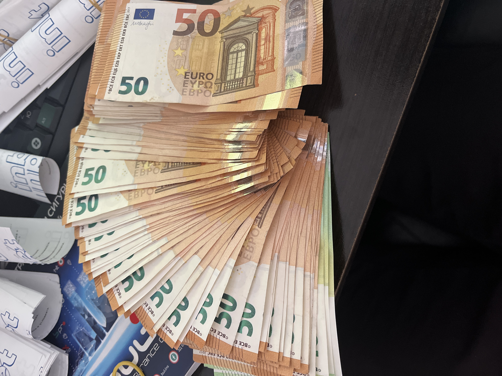 A substantial, fanned-out stack of 50 Euro banknotes is prominently displayed on a dark surface, likely a desk or tabletop. The banknotes are neatly arranged, showcasing their value and quantity. The visible text on the Euro notes includes "50", "EURO", "ЕВРО", and the signature "m. Draghi," along with the copyright year "2017," indicating they are from a recent series.
To the left of the money, several white paper rolls with blue text are visible. Some rolls appear secured with rubber bands. Text on these rolls includes "inl" and "in," and partially visible Cyrillic script such as "СИГУРИ" (Syguri, meaning 'insurance' or 'security' in Bulgarian).
Beneath the banknotes and paper rolls, various blue and white cards or documents are discernible. These documents feature English text like "surance" and "bul.ins," along with Bulgarian text such as "ЗД Б" and a Facebook logo, suggesting materials from a Bulgarian insurance company, possibly "Bul.ins" or a similar entity. The presence of "OFFER 2017" hints at promotional or policy documents.
The scene, set in Ivailo, Bulgaria, depicts a collection of financial materials – cash and business documents – suggesting a business or personal finance context, possibly an insurance office. No people or specific activities are shown, conveying a sense of order and readiness.