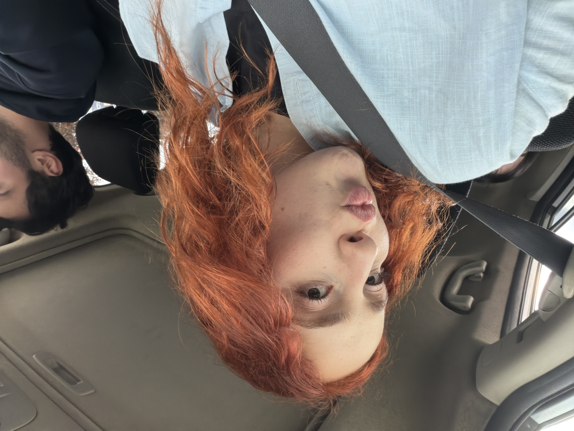 An upright, close-up image shows a woman and a man inside a car during daytime in Tbilisi, Georgia. The main subject, a woman, is positioned prominently on the right side of the frame. She has voluminous, wavy reddish-orange hair and fair skin with visible freckles. Her dark brown eyes gaze directly at the camera, and her lips are slightly pursed in a pouting or kiss-blowing expression. She is wearing a light blue, textured collared shirt over a darker garment, with a black seatbelt diagonally crossing her chest.
Behind her and to the left, the profile of a man is partially visible, showing his dark hair and beard. He is also seated in the car, wearing a dark top. The setting is clearly a car interior, with visible elements including car seats, headrests, a light-colored roof liner, a sun visor, and a grab handle. Bright lighting suggests it is daytime. The image appears to be a selfie or a closely framed portrait, taken from a low angle, capturing an engaging and playful moment. No text is visible in the image.