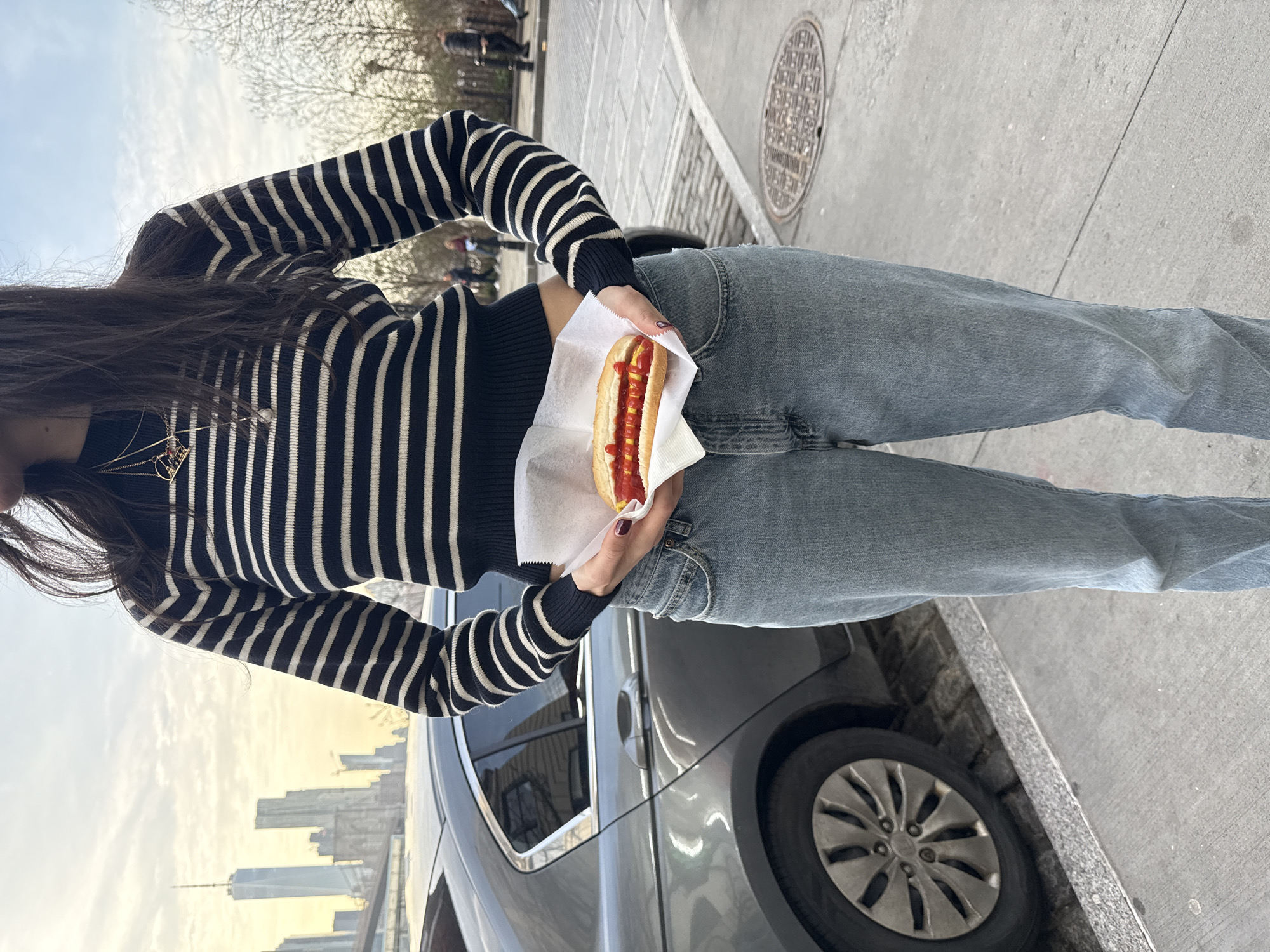 A woman stands on a sidewalk in New York City, holding a hot dog in a paper wrapper. She is wearing a black and white striped long-sleeve shirt and light blue jeans. The skyline of Lower Manhattan, including One World Trade Center, is visible in the background, and a car is parked nearby. A manhole cover is on the sidewalk.
