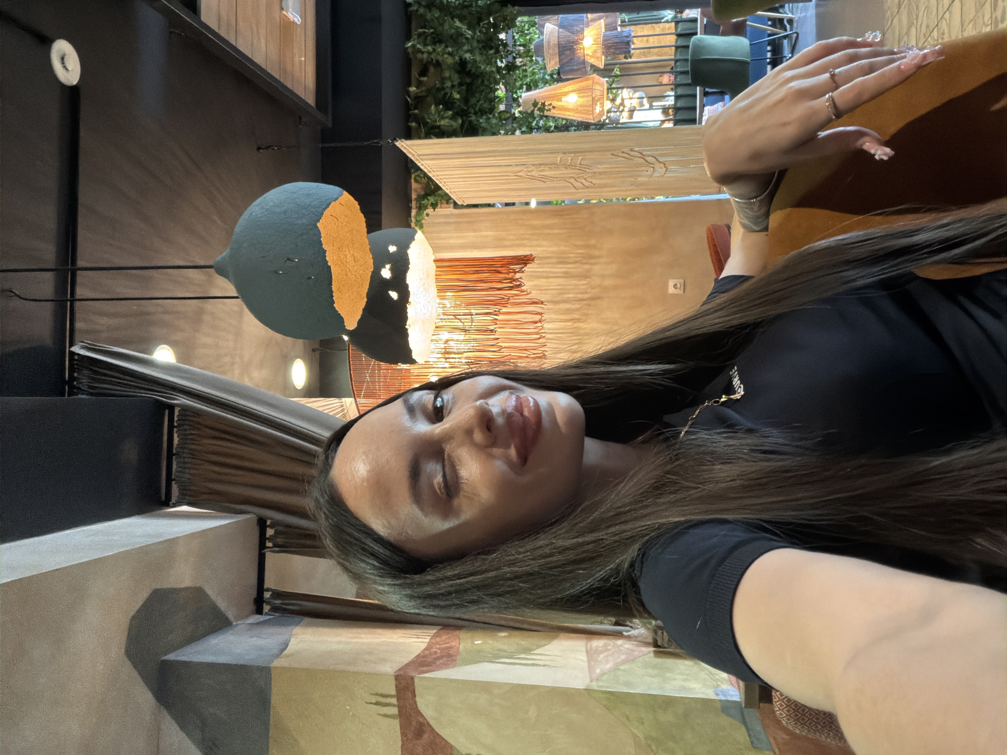 This media file is a selfie of a young woman, captured in a portrait orientation but rotated 90 degrees clockwise. The woman, with long, straight dark brown hair, occupies the right side of the frame. She is wearing a black top with partially visible white text that appears to say "URBAN" or "URBANE." Her face is turned towards the camera, her lips slightly parted, and her right eye is closed in what seems to be a wink or squint. Her left hand is visible in the upper right, resting on a brown surface, showcasing long, manicured nails and rings on her ring and middle fingers.
The background reveals the interior of what appears to be a stylish restaurant or cafe in Izhevsk, Russia. Various decorative light fixtures are prominent. To the left of the woman, a large, spherical pendant light is visible, dark teal/blue with a segment of bright orange. Further back, other light fixtures, some made of woven natural materials, hang amidst lush green foliage. The setting includes sections of walls with abstract, geometric patterns in shades of brown, beige, and green. A dark table or counter is also partially visible. The overall ambiance is indoors, likely evening or a dimly lit setting, suggested by the reliance on artificial lighting.