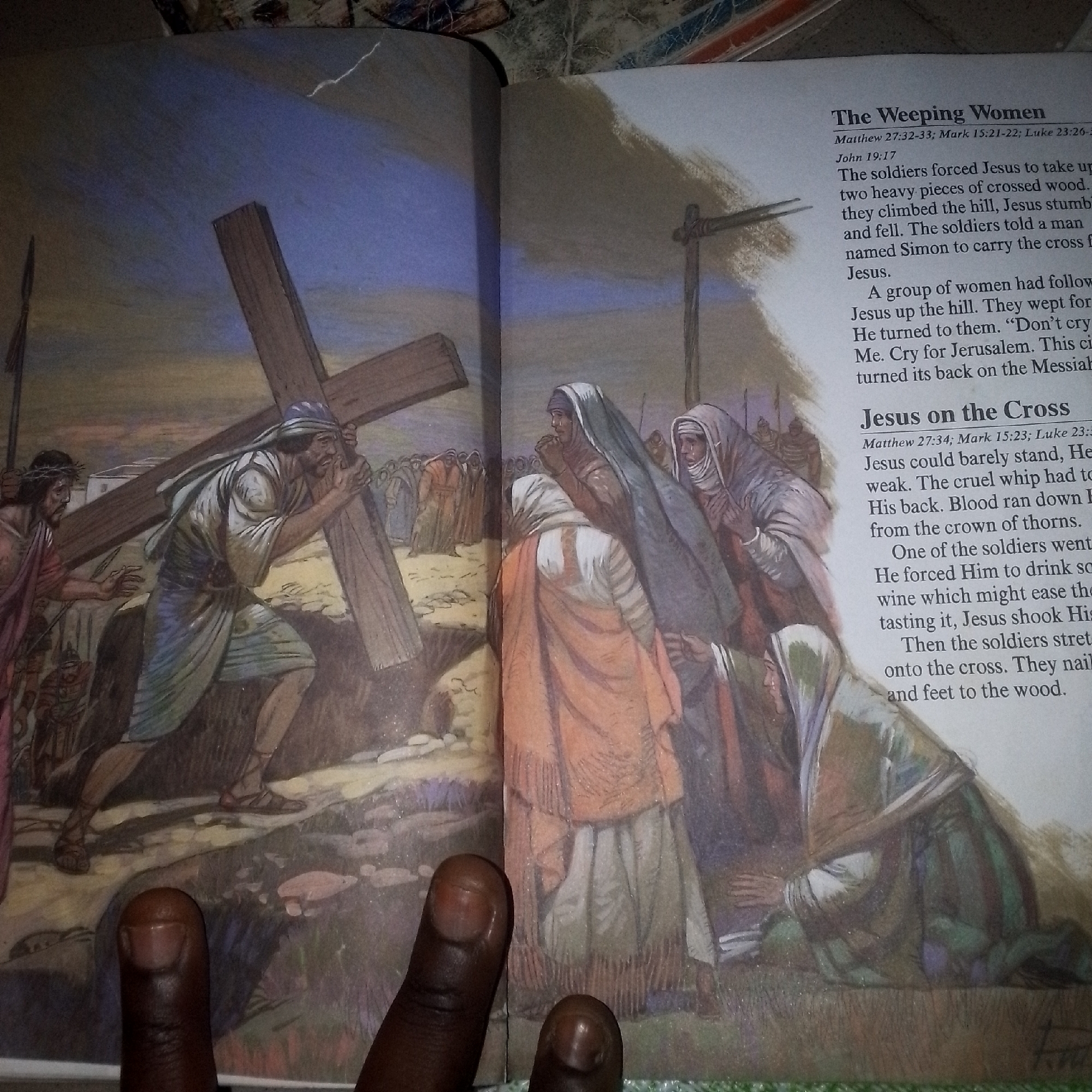 The image shows an open book with an illustration and text on both pages. The illustration depicts a scene from the Bible, likely the carrying of the cross by Jesus.
On the left page, a man wearing a purple robe and a crown of thorns, presumably Jesus, is shown walking. Behind him, a man named Simon is carrying a large wooden cross. They are on a path with other figures, some appearing to be soldiers, and a crowd of people in the background. The environment appears to be outdoors, possibly a hillside, with a barren landscape.
The right page contains text titled "The Weeping Women" and "Jesus on the Cross." The text describes the events of Jesus' journey to crucifixion, including Simon carrying the cross and Jesus' interaction with weeping women. The illustration on the right page shows a group of women in traditional attire, some with their heads covered, looking distressed and weeping. One woman is kneeling. The background on this page is less detailed, suggesting a more focused depiction of the women's grief.
The image is partially obscured by a hand with dark skin, with two fingers visible in the foreground, near the bottom of the book. The lighting in the image is not uniform, with some areas appearing brighter than others. The book appears to be an old edition, possibly a Bible, given the content.