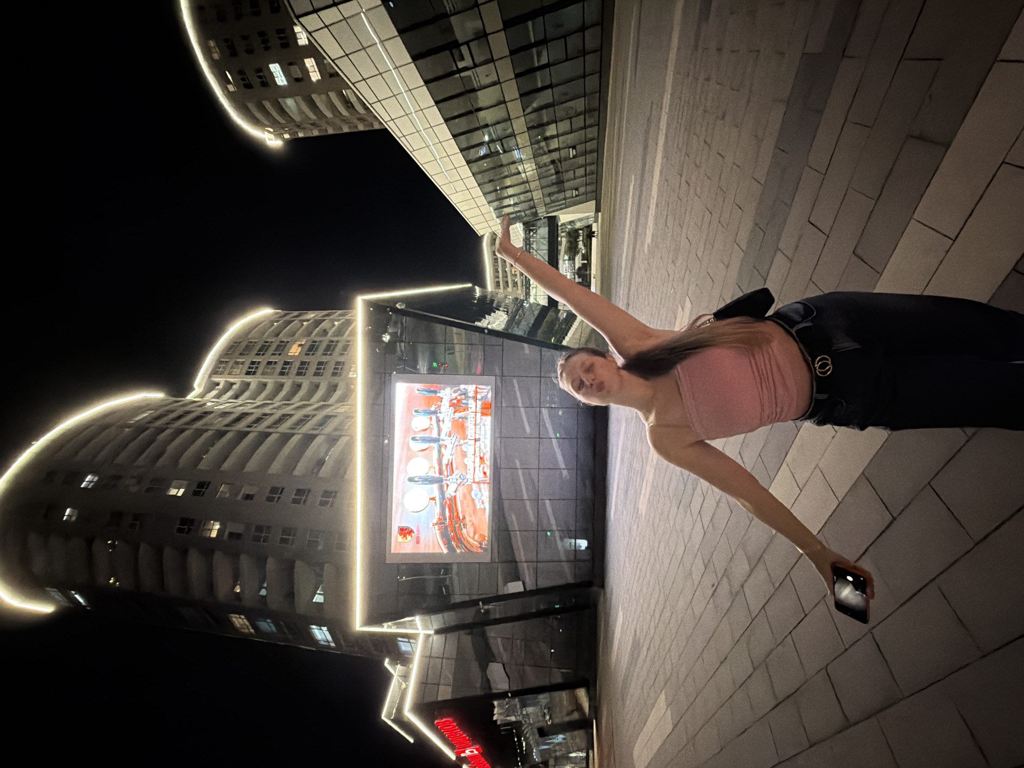 The image displays a young woman posing at night in a modern urban setting, identified as Burgas, Bulgaria. She stands on a paved ground made of light-colored, rectangular tiles. The woman has long, dark hair and is wearing a pink strapless top, dark trousers, and a dark belt with a gold-colored, double-ring buckle. Her left arm is raised with an open hand, while her right hand holds a dark smartphone pointed downwards. She looks directly at the camera with a slight smile, conveying a confident and cheerful demeanor.
In the background, two contemporary buildings with distinctive curvilinear architecture rise against the dark night sky. These structures feature extensive glass facades and are illuminated by bright white linear light strips tracing their edges.
On the building directly behind the woman, a large, brightly lit digital screen displays an advertisement. The ad primarily features several silver-colored beer taps or similar bar equipment, accompanied by reddish-orange elements, possibly depicting branding or drinks. Below this large screen, a smaller, horizontally oriented sign with red Cyrillic text is partially visible. The text begins with "ХРАНИТЕЛНИ...", which translates to "FOOD..." in Bulgarian, indicating a food-related establishment like a grocery store. The entire scene is brightly illuminated by the building's architectural lights and advertising displays, creating a vibrant night atmosphere.