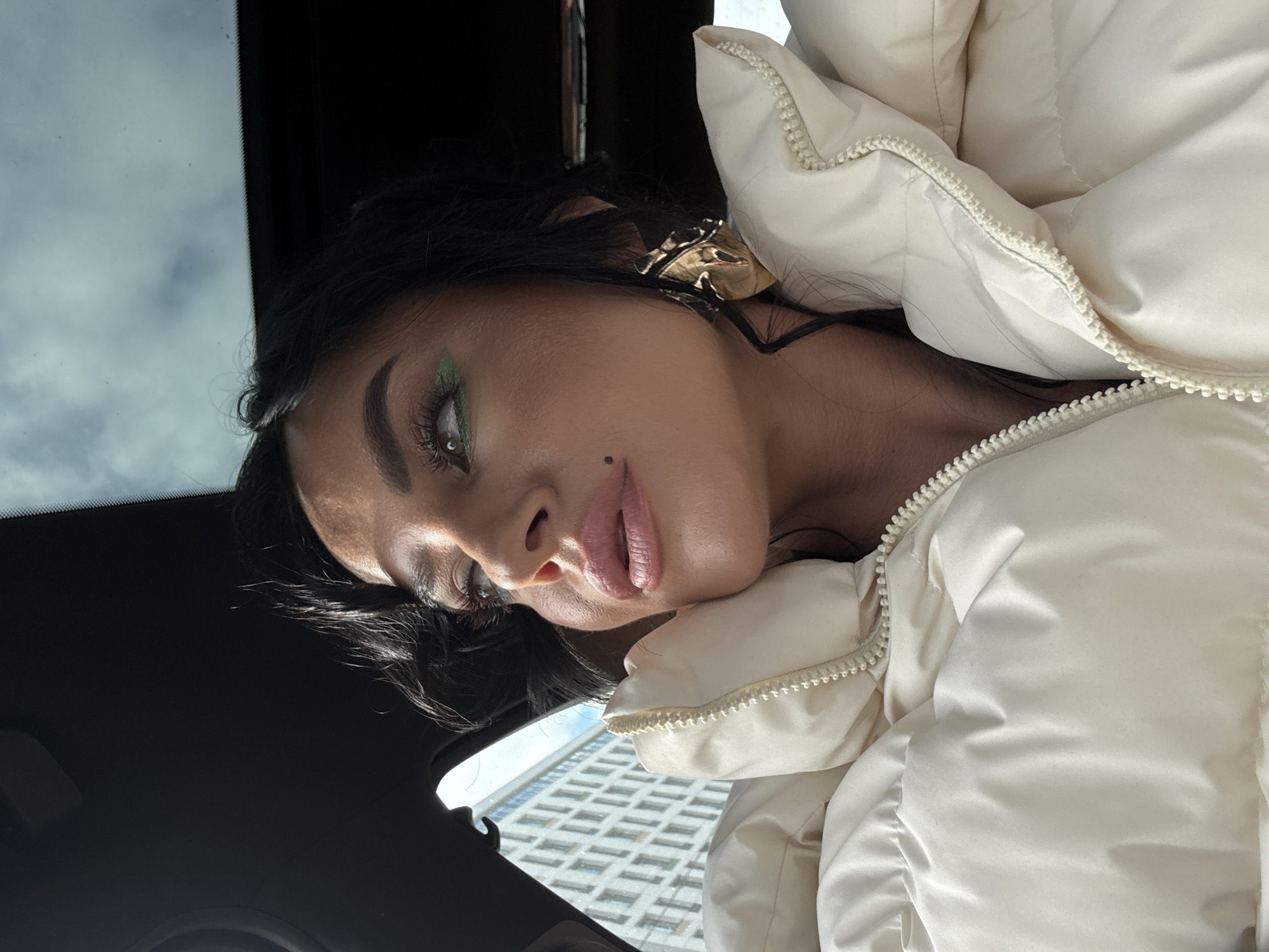 The media file displays a close-up, chest-up portrait of a woman, taken from a slightly low angle, inside what appears to be a car in Krasnoyarsk, Russia. The woman's head is tilted to her right, and she looks directly towards the camera with a neutral to slightly alluring expression and lightly parted lips.
She has dark, wavy hair. Her makeup is prominent, featuring strong dark eyeliner and a vibrant line of bright green eyeshadow along her lower lash line. She appears to be wearing false eyelashes, with a small reflective jewel or glitter near the inner corner of her left eye. A beauty mark or mole is visible below her right nostril, and her lips are a soft pink.
She is dressed in a thick, light-colored (cream or off-white) puffy winter jacket, which has a visible zipper running down its front. A large, abstract, golden earring is visible in her left ear.
The background indicates she is inside a vehicle. Above and behind her, the dark interior roof of a car is visible. Through the car's windshield, a cloudy sky can be seen in the upper left portion of the background, while a tall building with a distinctive grid pattern of numerous windows is visible in the lower right, suggesting an urban environment. The lighting is natural and bright, consistent with daytime, casting subtle highlights and shadows. No text is visible in the image.