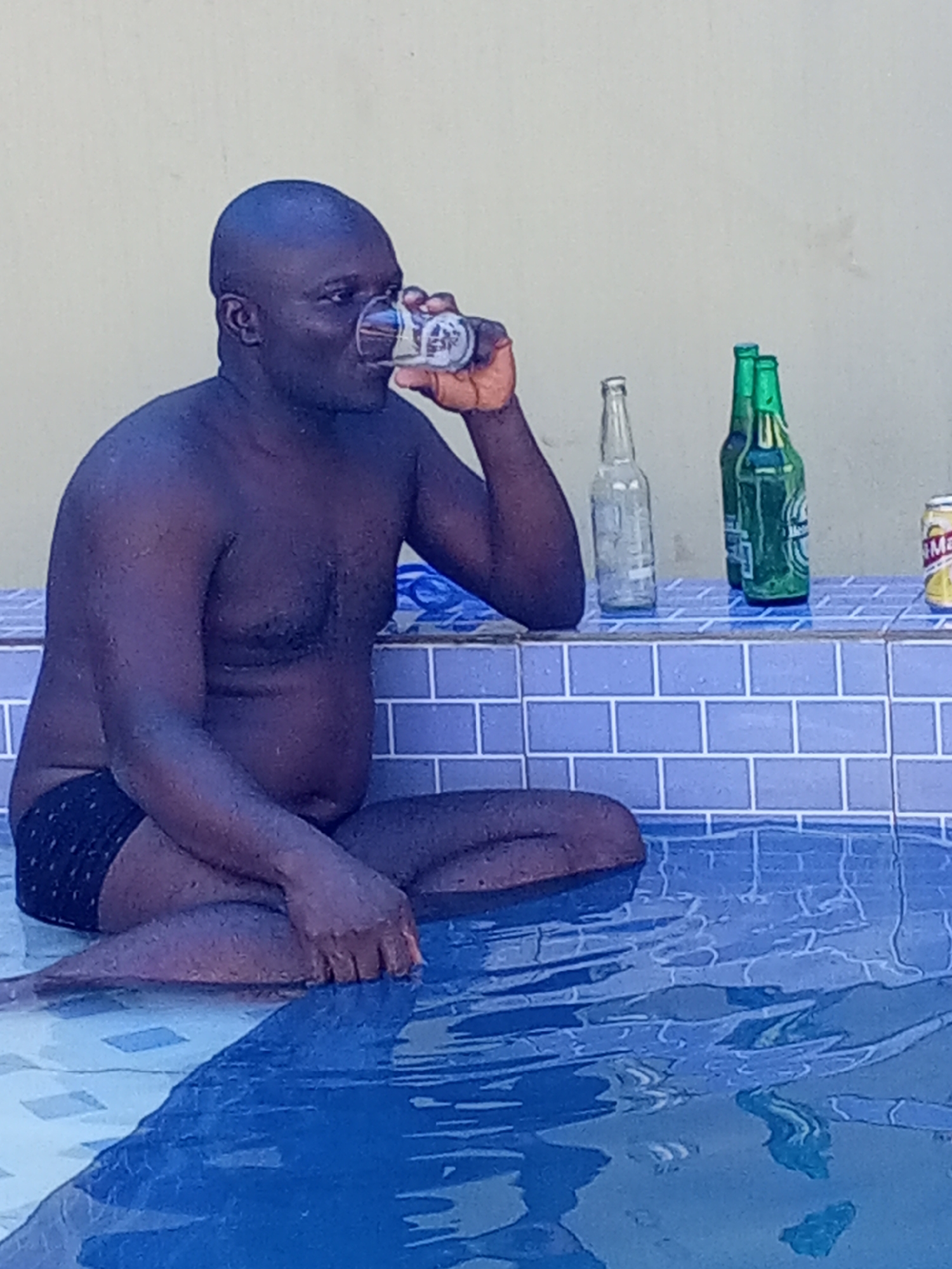 A shirtless, dark-skinned man is sitting cross-legged in a tiled swimming pool, drinking from a glass. He is wearing dark swim trunks. On the tiled pool ledge behind him are an empty glass bottle, two green beer bottles, and a yellow beverage can. The location is likely Ankpa, Nigeria.