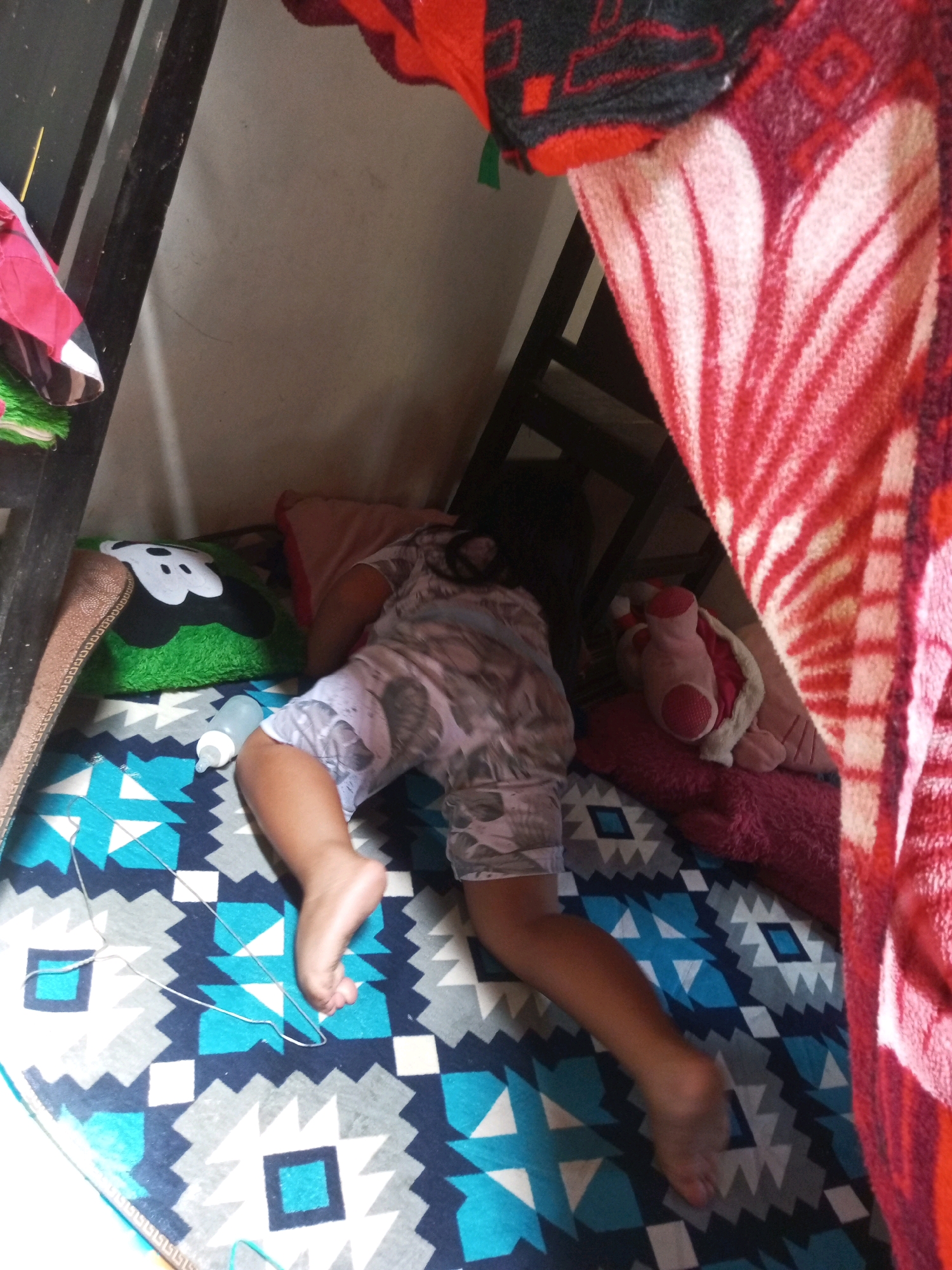 A child sleeps on a colorful, patterned blanket with a Mickey Mouse pillow beside them. A baby bottle is also visible on the blanket. The scene appears to be in a bedroom.