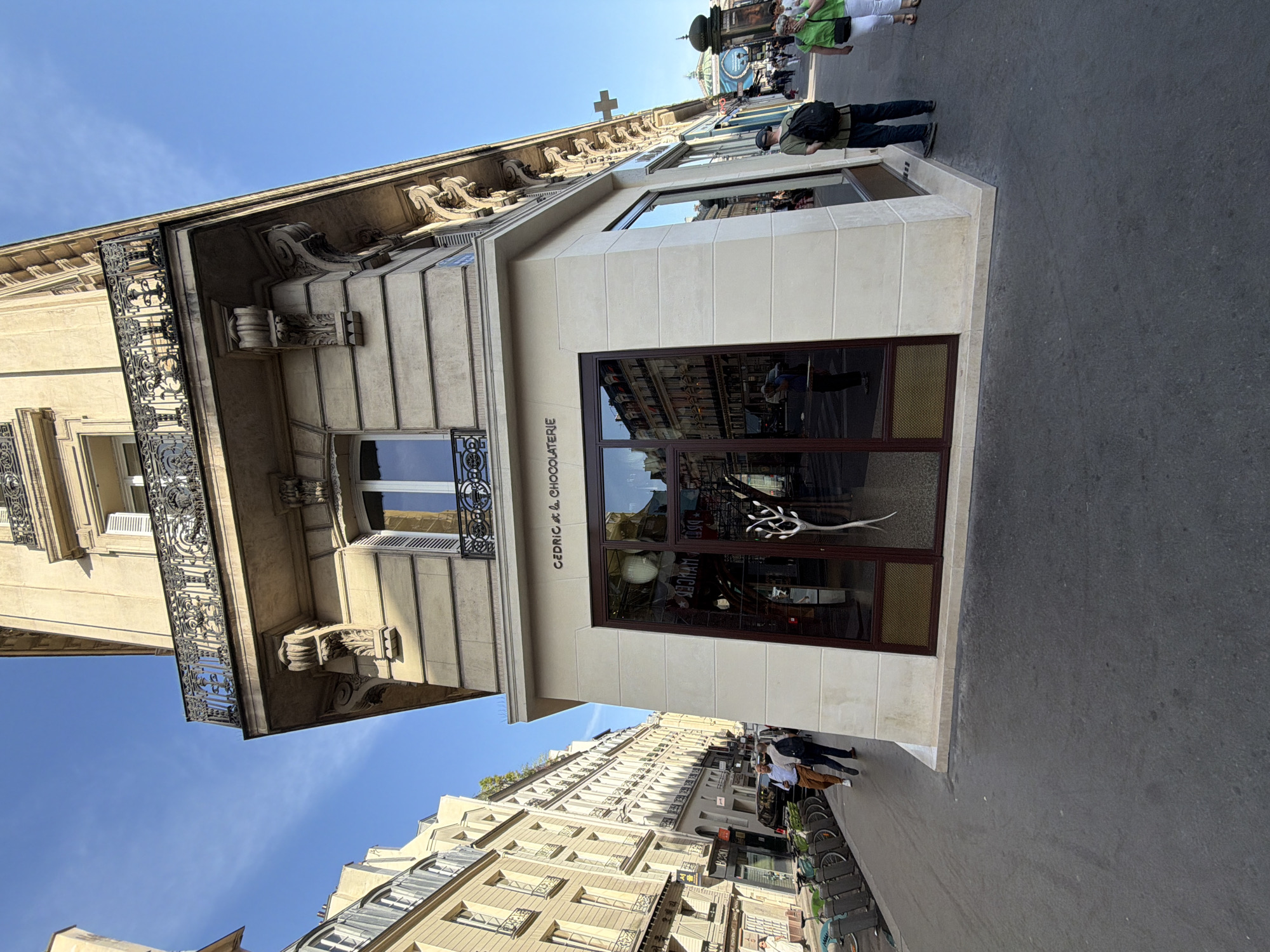 The image depicts a street scene in Paris, France, featuring the storefront of "Cedric Grolet Chocolaterie." The building has a classic Parisian architectural style with ornate details and a stone facade. The storefront is modern, with large glass windows displaying interior elements. People are walking along the sidewalk in front of the shop. The sky is blue, suggesting a sunny day. Other buildings line the street, showcasing typical Parisian architecture.