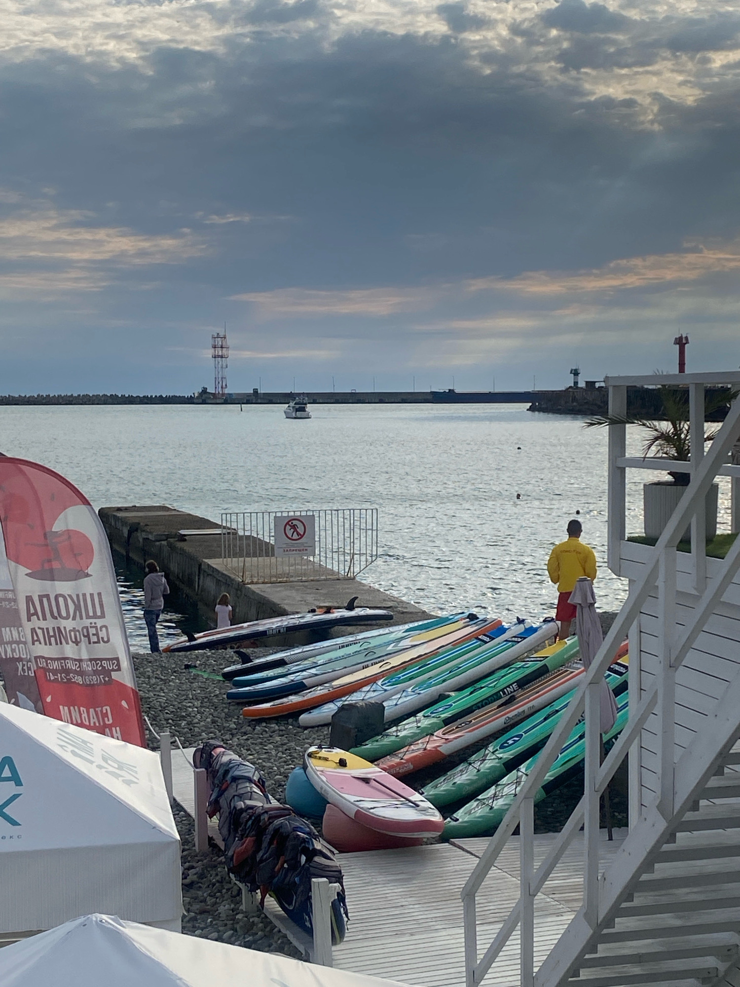 The image depicts a cloudy day at a beach, likely in Sochi, Russia, given the presence of a surf school banner and the distinctive coastal structures. In the foreground, a large red and white banner with Russian text that appears to advertise a surf school is partially visible. Next to it, several paddleboards are lined up on a pebble beach. A white staircase with handrails ascends on the right side of the frame. In the middle ground, two people, an adult and a child, stand on a concrete pier that extends into the calm, gray water. A white boat is visible in the distance, navigating the sea towards a long, man-made breakwater. Along the breakwater, there are industrial structures, including a tall red and white transmission tower and a red lighthouse. A person wearing a yellow jacket and red shorts, possibly a lifeguard, stands near the water's edge, facing away from the camera. The sky is overcast with a mix of dark and lighter clouds, suggesting either early morning or late afternoon. The overall scene conveys a sense of preparedness for water activities, despite the less-than-ideal weather.