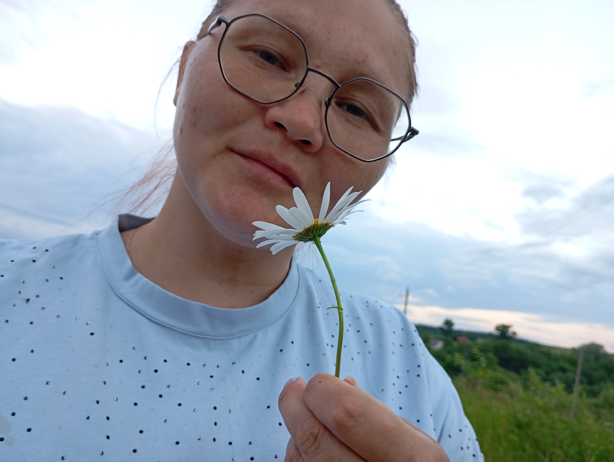 A medium shot shows a person wearing glasses and a light blue shirt with scattered dark and shiny embellishments. The person is holding a single white daisy with a yellow center near their face, with the flower positioned in front of their lips. The background is a hazy outdoor scene with a pale, cloudy sky and a hint of green foliage and distant structures on a hillside. The image does not contain any information about the location in the city of Ryabki, Russia.