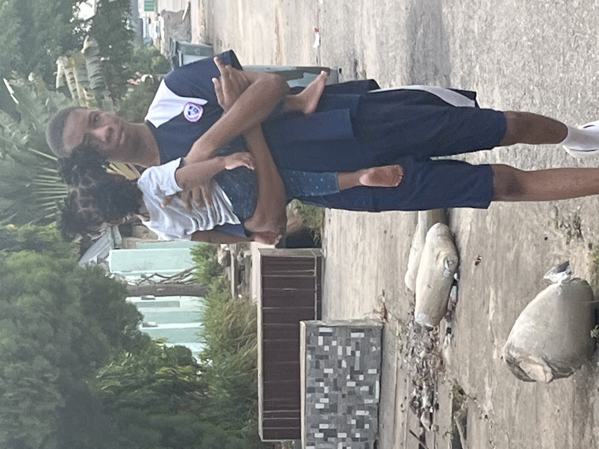 An adult male is pictured outdoors carrying a child, both facing the viewer. The image is taken in daylight under what appears to be an overcast sky.
The adult, a male with dark skin and short dark hair, is wearing a dark blue and white short-sleeved t-shirt with a purple, white, and blue circular emblem on the left chest. He also wears dark blue shorts, white socks, and white sneakers. He is holding the child in a front carry position, with the child's arms around his neck and legs around his waist. He has a neutral expression looking towards the camera.
The child, with dark skin and braided hair, is largely obscured by the adult's body, but is wearing a white t-shirt and dark blue pants or shorts with light-colored patterns. Their bare feet are visible, one dangling freely and the other resting against the adult's leg.
The setting appears to be a paved or concrete outdoor area, possibly a pathway or yard. To the left and in the background, there is abundant green foliage from trees and bushes, suggesting a tropical environment consistent with Abuja, Nigeria. In the middle ground, a dark brown rectangular structure and a grey and white tiled block are visible. Further in the background, light-colored buildings can be seen through the trees. Several off-white, sack-like objects or large rocks are scattered on the concrete ground to the right.
No explicit text is clearly legible, though there is a logo on the adult's shirt that cannot be deciphered. The overall mood is casual and familial.