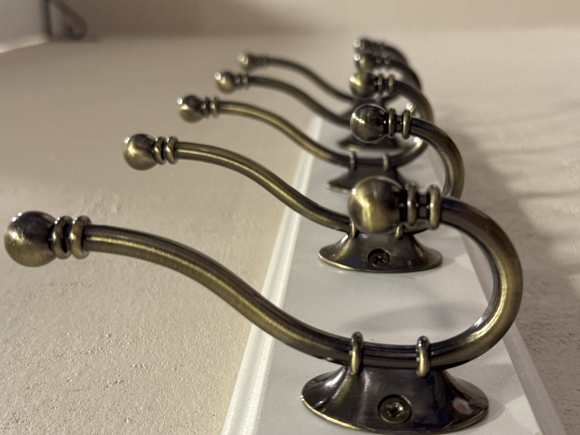 The image displays a row of antique brass coat hooks mounted on a white wooden strip. The hooks are arranged in a linear fashion and are partially obscured by a shallow depth of field, with the closest hook in sharp focus and the subsequent hooks becoming progressively blurred. Each hook has a rounded finial at its tip and a decorative beaded detail on its shaft. The hooks are attached to the white strip with small screws, visible on the base of each hook. The background is a textured, off-white wall. The lighting suggests an indoor setting, possibly a home or a retail display. There are no people or animals visible in the image, and no text is present.