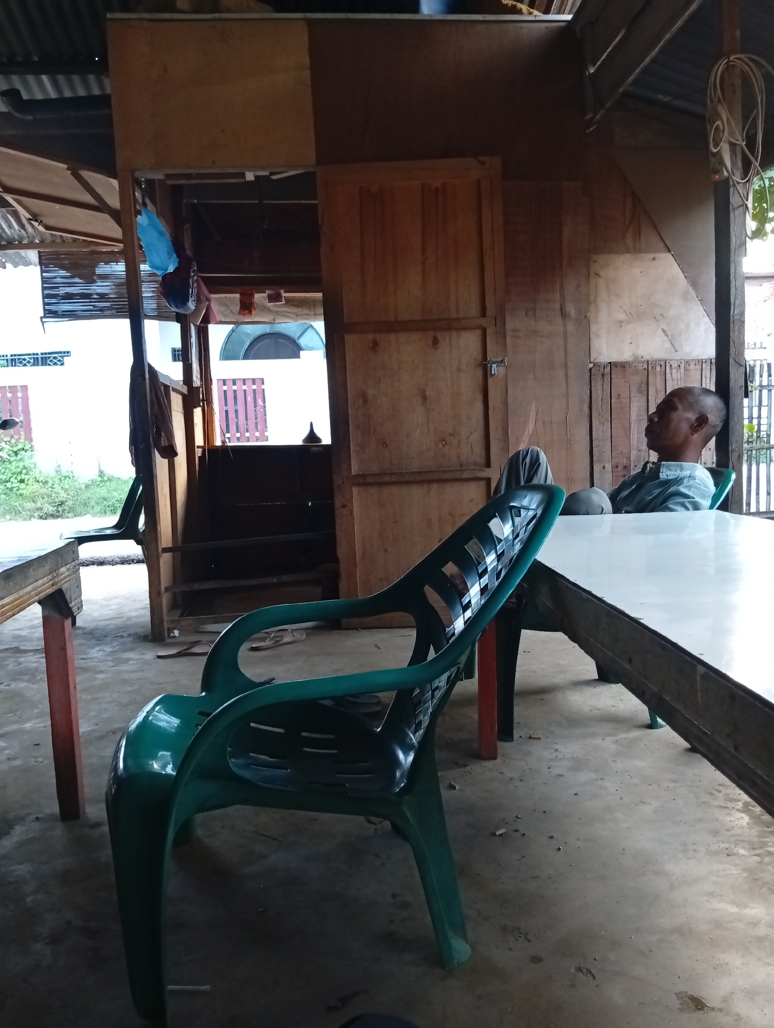 The image depicts an outdoor seating area, likely a small food stall or rest stop, in Lhokseumawe, Indonesia. A middle-aged man of Asian descent, wearing a light blue collared shirt and grey pants, is seated in a dark green plastic chair, leaning back with his head resting against the chair's back. He appears to be looking towards the right, his facial expression neutral. In the foreground, another dark green plastic chair is positioned at an angle. A long, rectangular table with a white or light-colored top and a weathered wooden frame separates the two chairs and the man. The table has a rough texture, suggesting it is made of wood. The surrounding environment is rustic, with unfinished wooden walls and a corrugated metal roof visible. The floor is concrete, showing signs of wear and some scattered debris. Behind the man, a doorway leads into a darker interior space. The overall impression is of a casual, informal gathering spot.