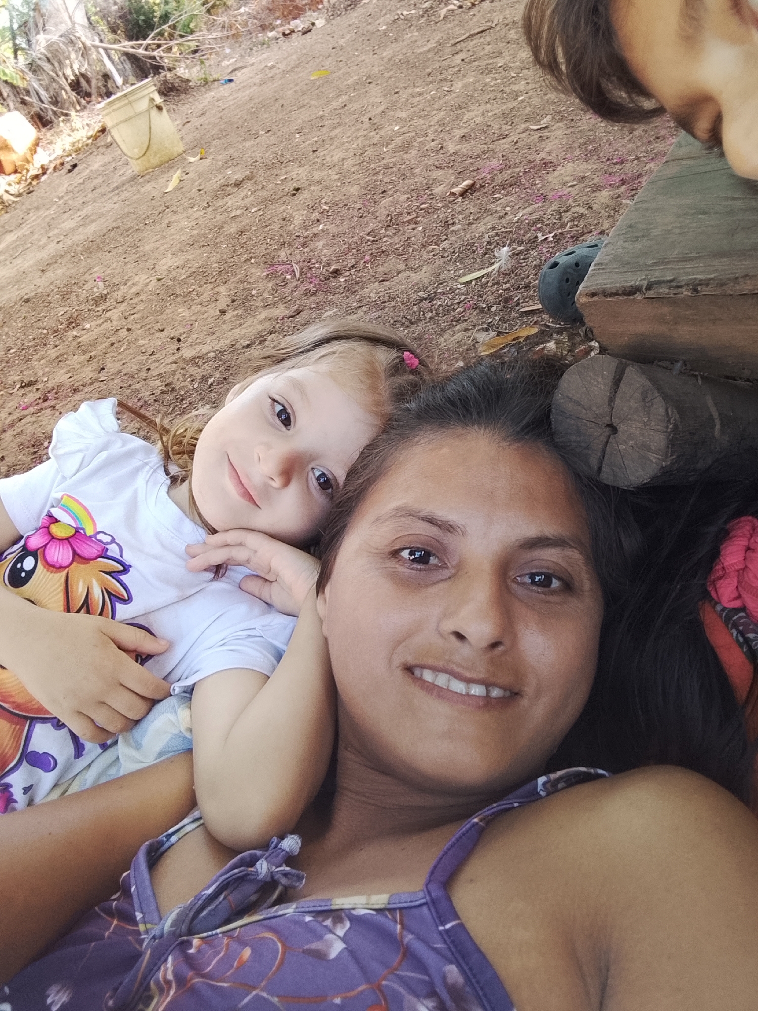 A selfie-style image shows a woman and a young child lying on the ground. The woman is facing the camera, smiling with dark hair. The child, to the left of the woman, is fair-skinned with light hair and also smiling. The background consists of bare earth, logs, and foliage. A bucket is visible in the upper left corner. A Croc shoe is on the upper right. While Altagracia, Venezuela is provided as information, the image does not display anything indicative of that.