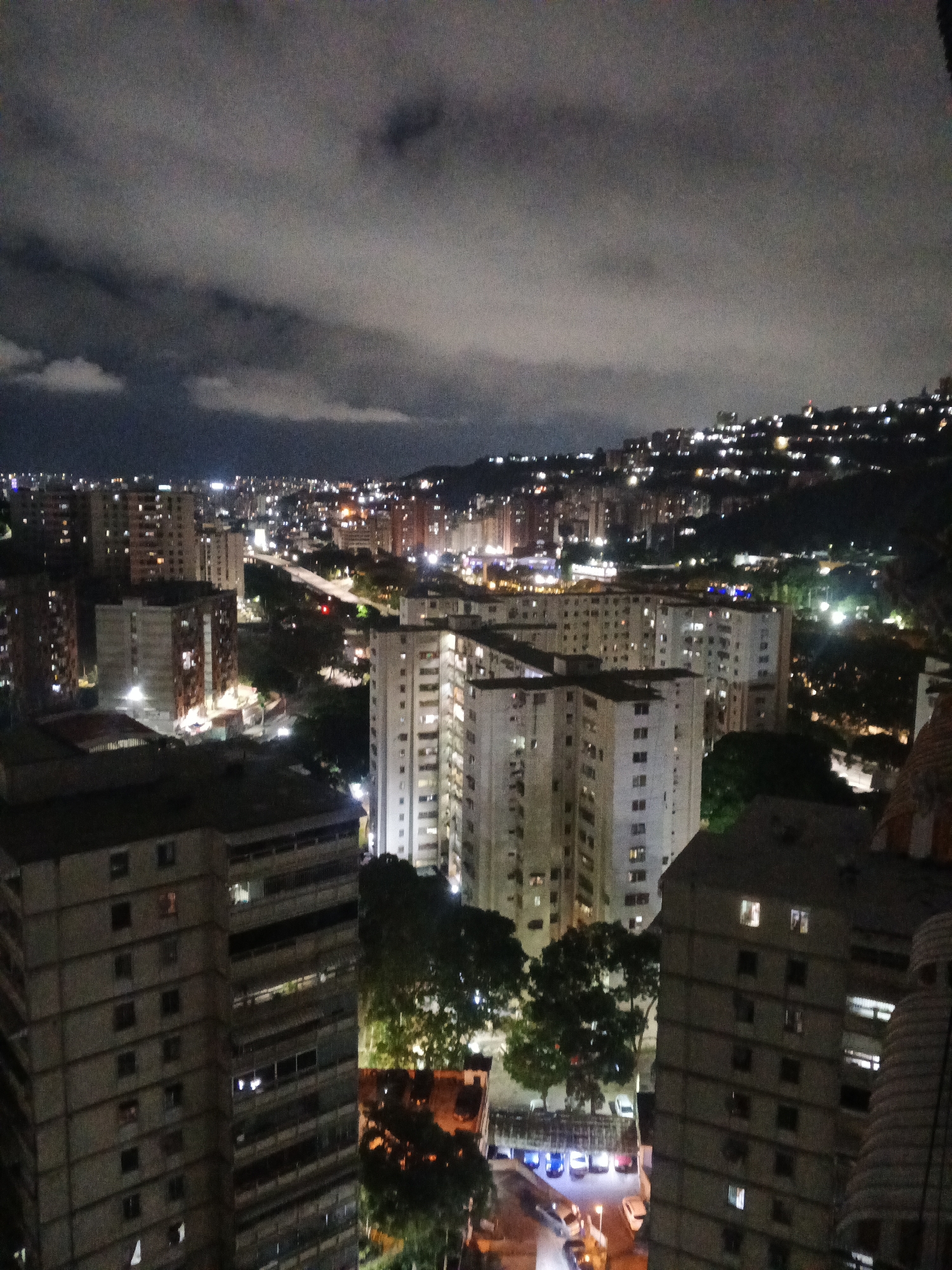 The image depicts a night view of Caracas, Venezuela. The scene is dominated by numerous high-rise apartment buildings, many illuminated with internal lighting. A highway overpass is visible in the middle distance, also lit. Further back, the landscape rises into hills, dotted with clusters of lights indicating residential areas. The sky is overcast with heavy, dark clouds. The scene appears calm and still, with no visible people or activity.